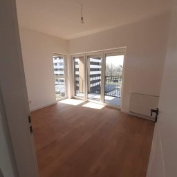 RM Produkcja - Jasne, puste wnętrze z parkietem i balkonem w nowym budownictwie. Świeżo pomalowane ściany, gotowe do umeblowania. Widok zza uchylonych drzwi.