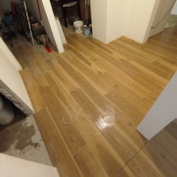 RM Produkcja - Ukończona podłoga z płytek imitujących drewno w korytarzu, z widocznymi wiaderkami i narzędziami w tle, sugerująca świeżo zakończone prace wykończeniowe.