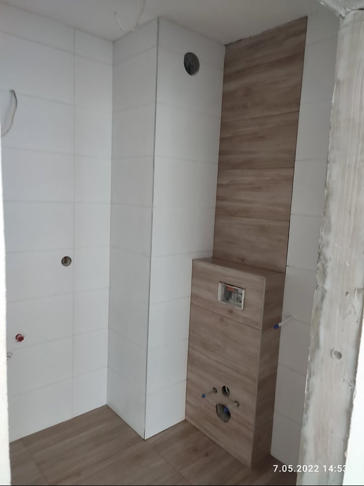 Białe i drewnopodobne płytki w łazience z zabudową WC. Widoczne otwory na armaturę i instalacje. Ukończony etap prac wykończeniowych w pomieszczeniu.