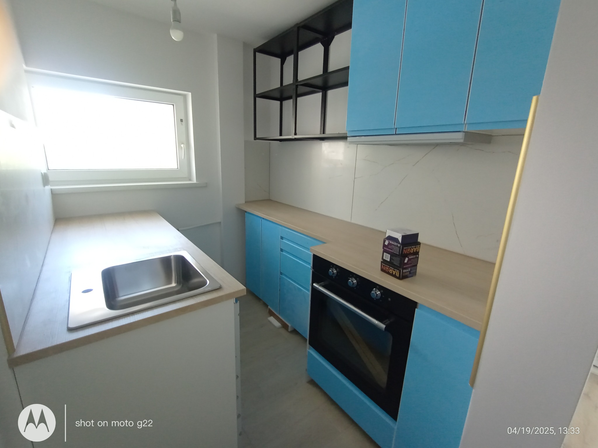 Niebieska zabudowa kuchenna z piekarnikiem i metalowym regałem na ścianie. Widok na jasne blaty i białe marmurowe płytki. Nowoczesne wykończenie kuchni.