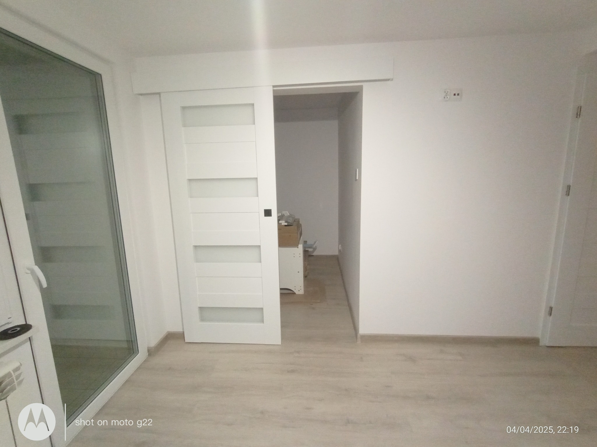 Białe, przesuwne drzwi z poziomymi przeszkleniami, zamontowane w jasnym wnętrzu. Widoczny fragment okna balkonowego i podłoga imitująca drewno.