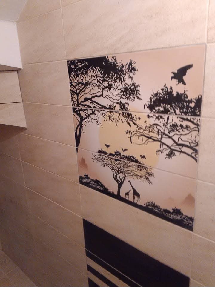 Beżowe płytki z czarnym motywem afrykańskim: drzewa, żyrafy i ptaki na tle zachodzącego słońca. Dekoracyjna ściana w łazience, widok z boku, lekki kąt.