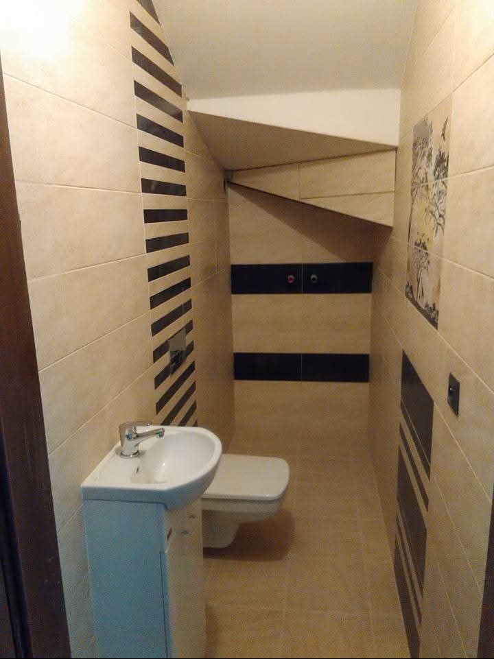 Wykończona łazienka w beżach z czarnymi akcentami. Umywalka z szafką, toaleta, płytki na ścianach i podłodze. Nietypowe rozwiązanie zabudowy nad toaletą.