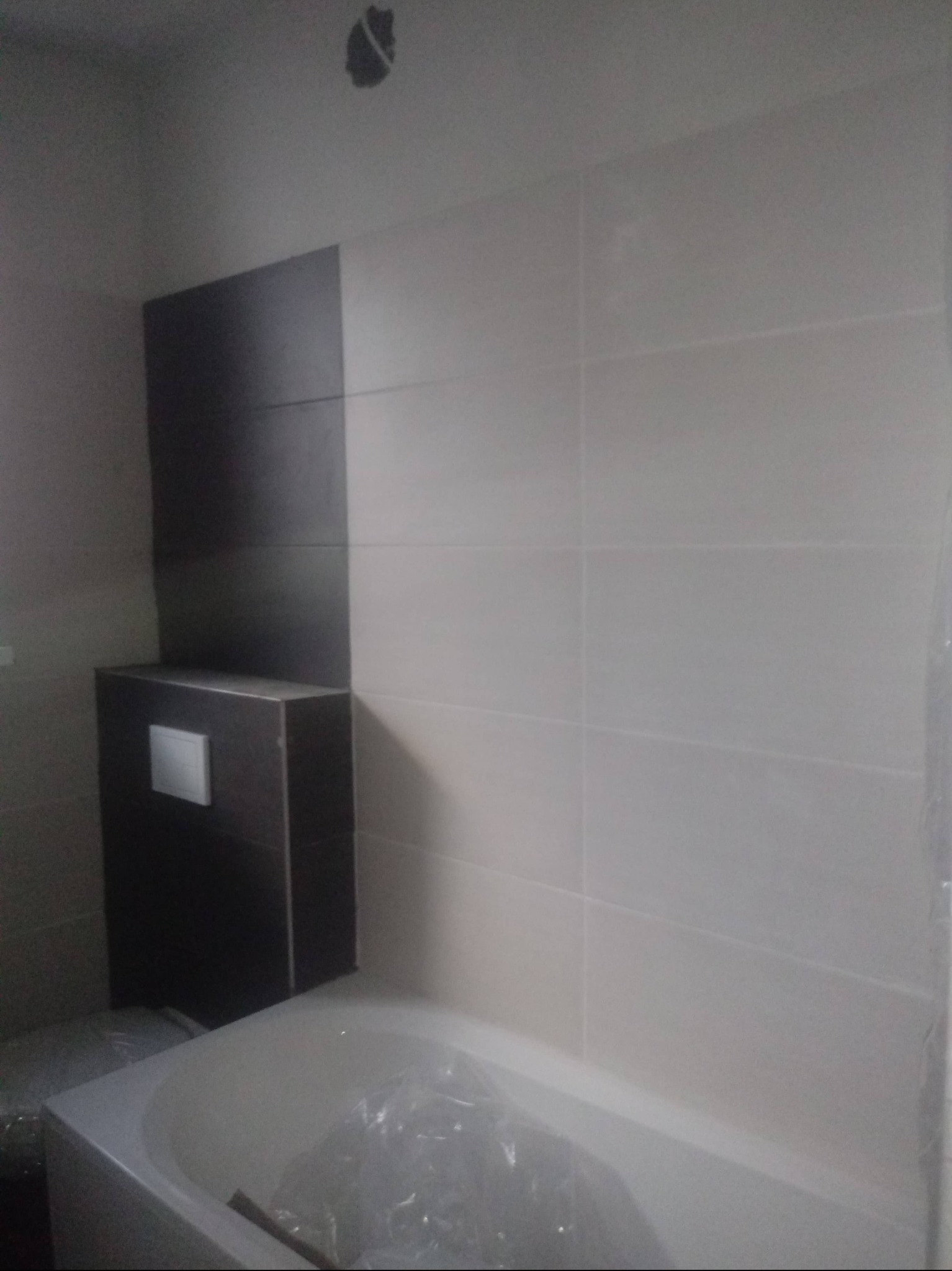 Wykończona łazienka z beżowymi płytkami ułożonymi poziomo i zabudową WC w kolorze grafitowym. Widoczna wanna zabezpieczona folią. Otwór w suficie.