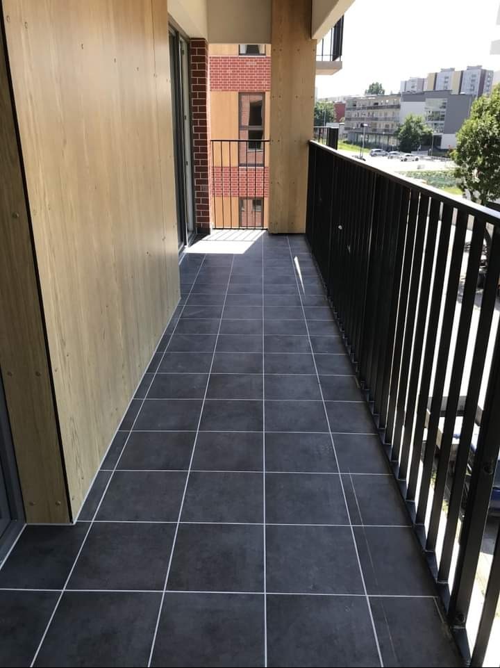 Wykończony balkon z ciemnymi, kwadratowymi płytkami i jasnymi fugami. Widok na zabudowę miejską. Nowoczesne balustrady i drewniane panele boczne.