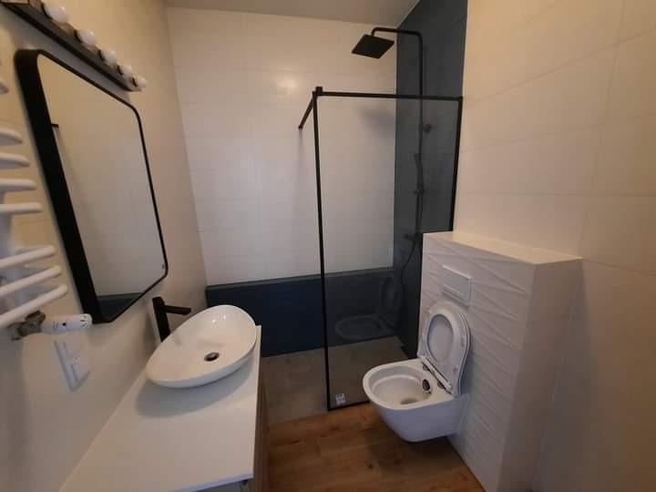 Nowoczesna łazienka z białą umywalką nablatową, czarną armaturą i kabiną prysznicową typu walk-in z ciemnymi płytkami w tle. Uwagę zwraca toaleta w zabudowie.