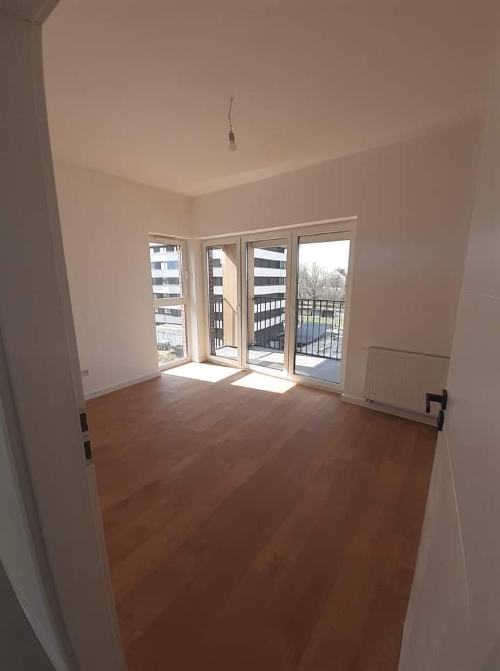 Jasne, puste wnętrze z parkietem i balkonem w nowym budownictwie. Świeżo pomalowane ściany, gotowe do umeblowania. Widok zza uchylonych drzwi.