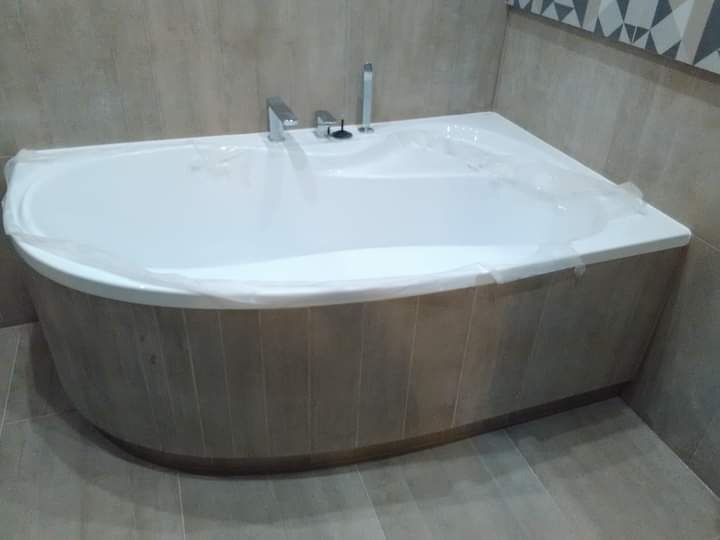 Narożna wanna z obudową imitującą drewno, zabezpieczona folią ochronną, w łazience wykończonej beżowymi płytkami i dekoracyjnym pasem na ścianie.