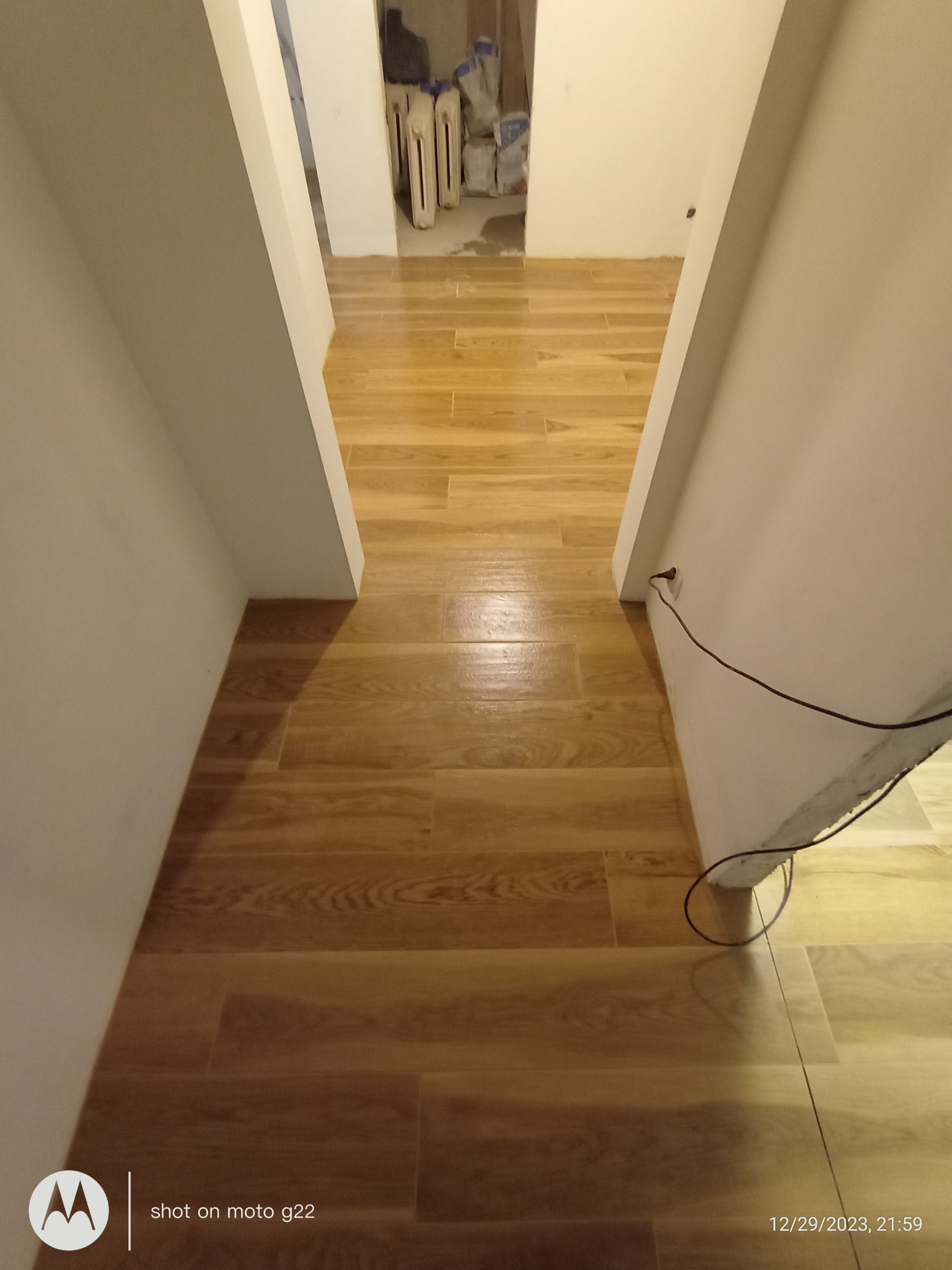 Ukończone układanie paneli podłogowych w korytarzu, widok z góry. Jasne, drewnopodobne panele ułożone równolegle do ścian, widoczne grzejniki w tle.