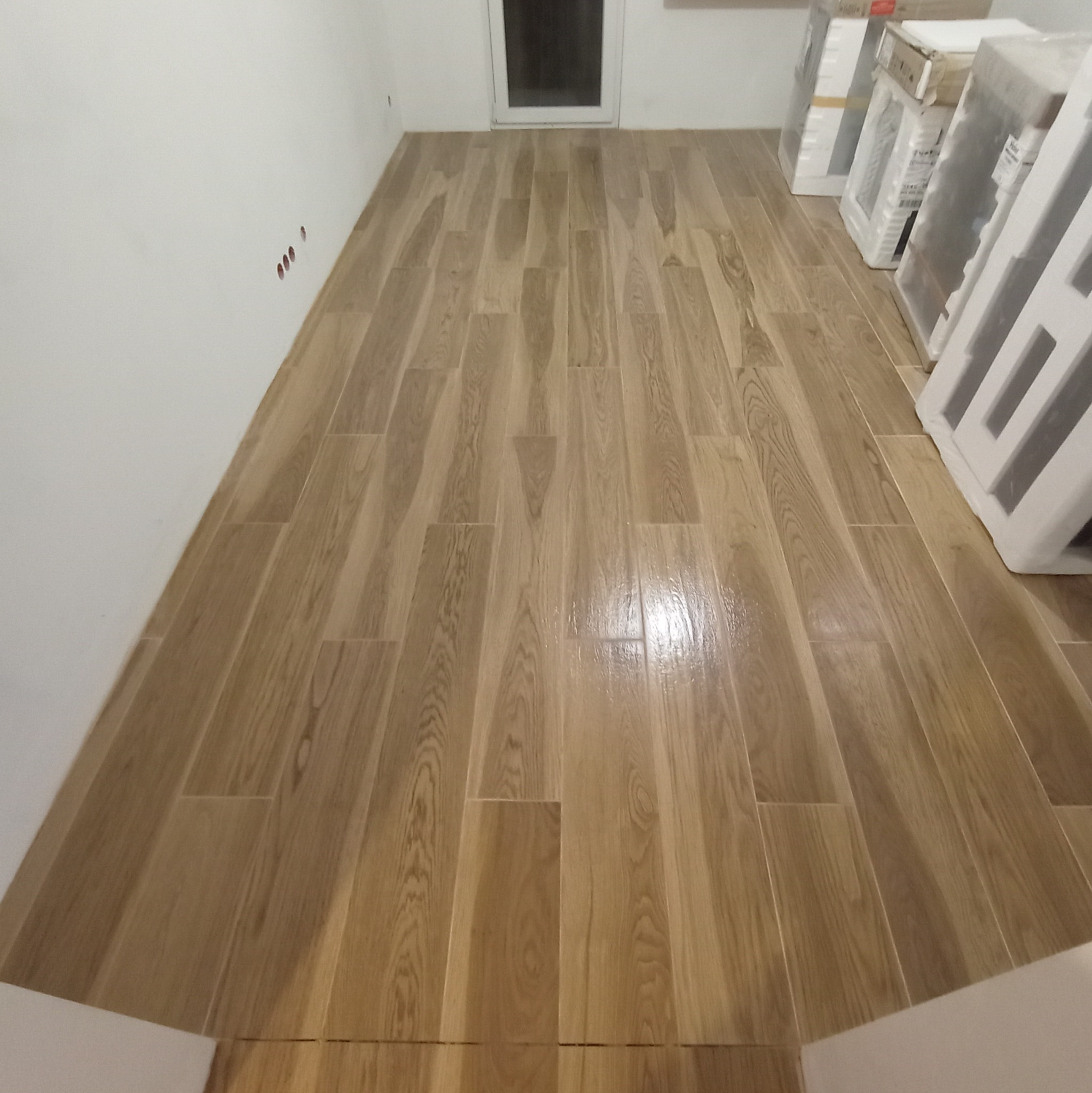 Ułożona podłoga z paneli imitujących drewno w jasnym odcieniu. Widoczne białe ściany i zapakowane materiały budowlane. Perspektywa z dołu podkreśla długość pomieszczenia.