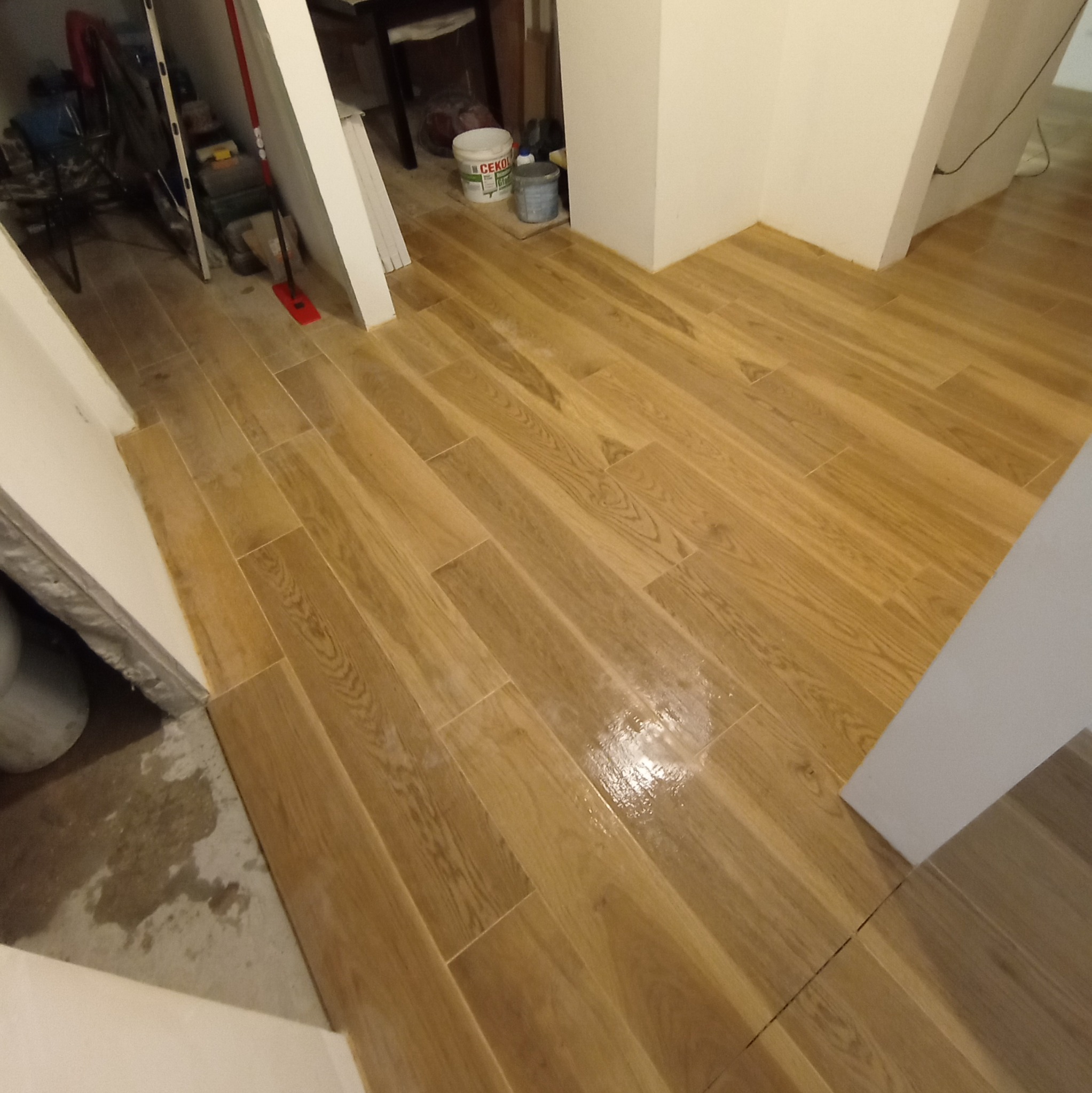 Ukończona podłoga z płytek imitujących drewno w korytarzu, z widocznymi wiaderkami i narzędziami w tle, sugerująca świeżo zakończone prace wykończeniowe.