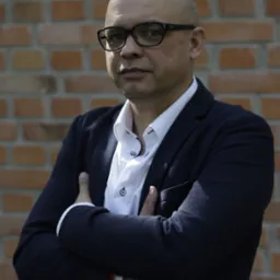 Portret pewnego mężczyzny w okularach, ubrany w białą koszulę i granatową marynarkę, stojącego na tle ceglanej ściany.