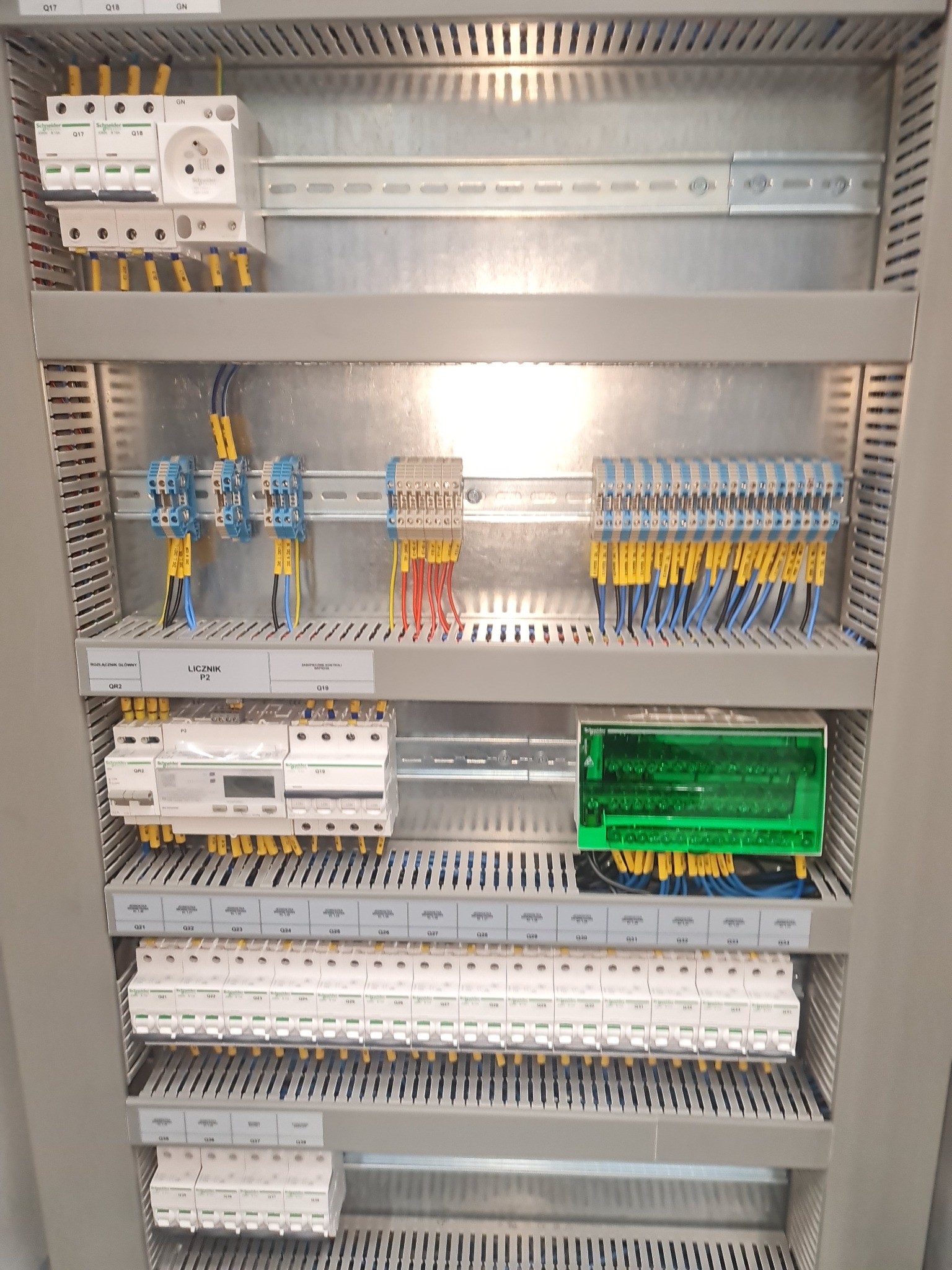 Rozdzielnica elektryczna z zamontowanymi bezpiecznikami, licznikiem i okablowaniem, widoczne oznaczenia na przewodach i etykiety opisujące elementy.