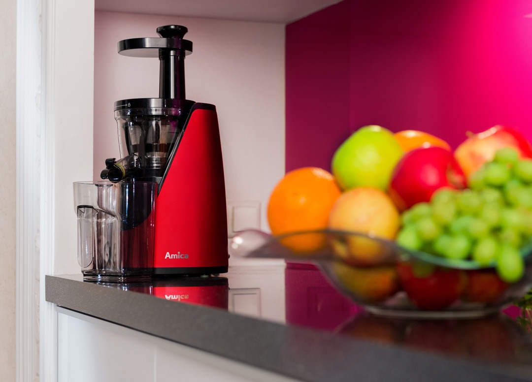 Czerwona wyciskarka wolnoobrotowa Amica stojąca na blacie obok szklanek i misy z owocami na tle ściany w kolorze fuksji.
