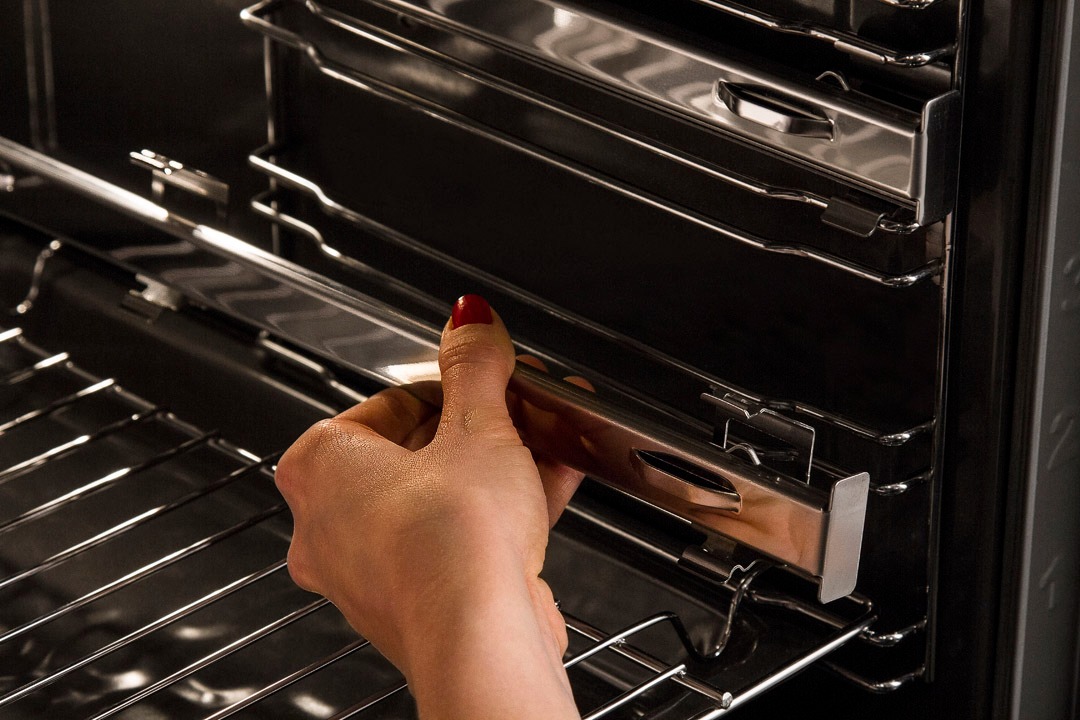 Wnętrze piekarnika z wysuwanymi prowadnicami teleskopowymi, dłoń z czerwonym paznokciem umieszcza prowadnicę na odpowiednim poziomie.