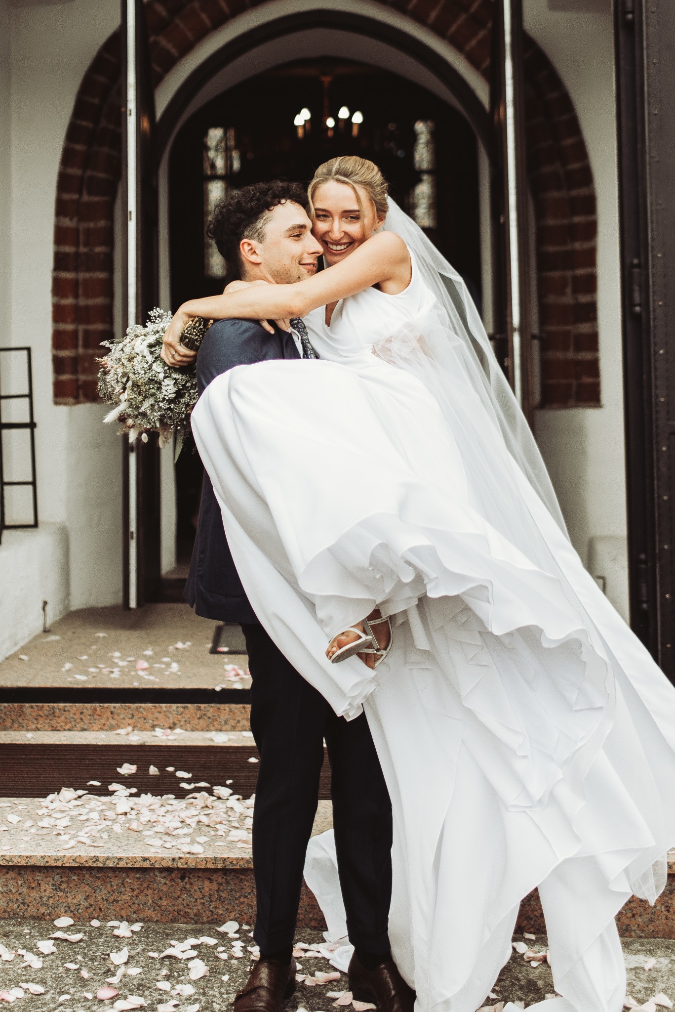 Uśmiechnięty pan młody niesie pannę młodą na rękach przed wejściem do budynku z ceglanymi łukami, biała suknia z welonem powiewa, bukiet w dłoniach mężczyzny, płatki róż na schodach.