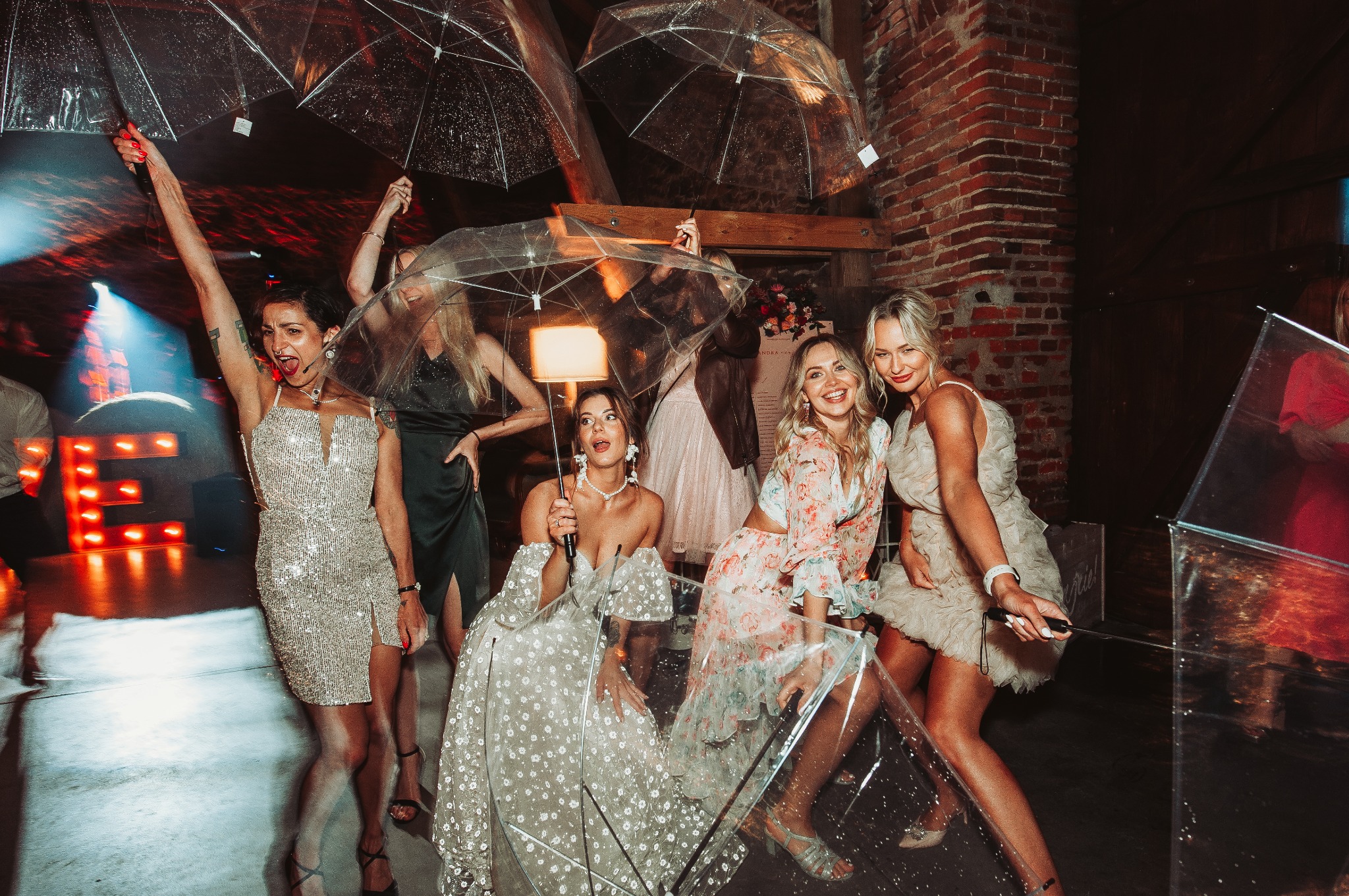 Grupa kobiet w wieczorowych kreacjach pozuje z przezroczystymi parasolami na sali z ceglanymi ścianami i dekoracjami kwiatowymi, oświetlona kolorowymi światłami, tworząc radosną atmosferę.