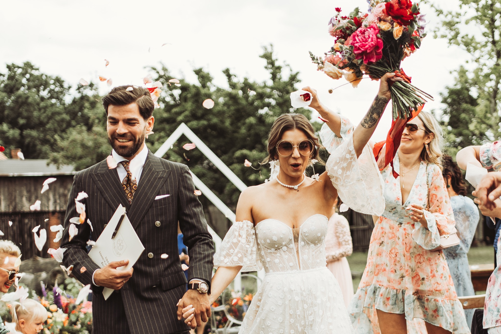 Para młoda wychodzi z ceremonii, obsypywana płatkami róż, panna młoda w sukni z koronkowymi rękawami, pan młody w eleganckim garniturze w prążki, goście weselni w tle.