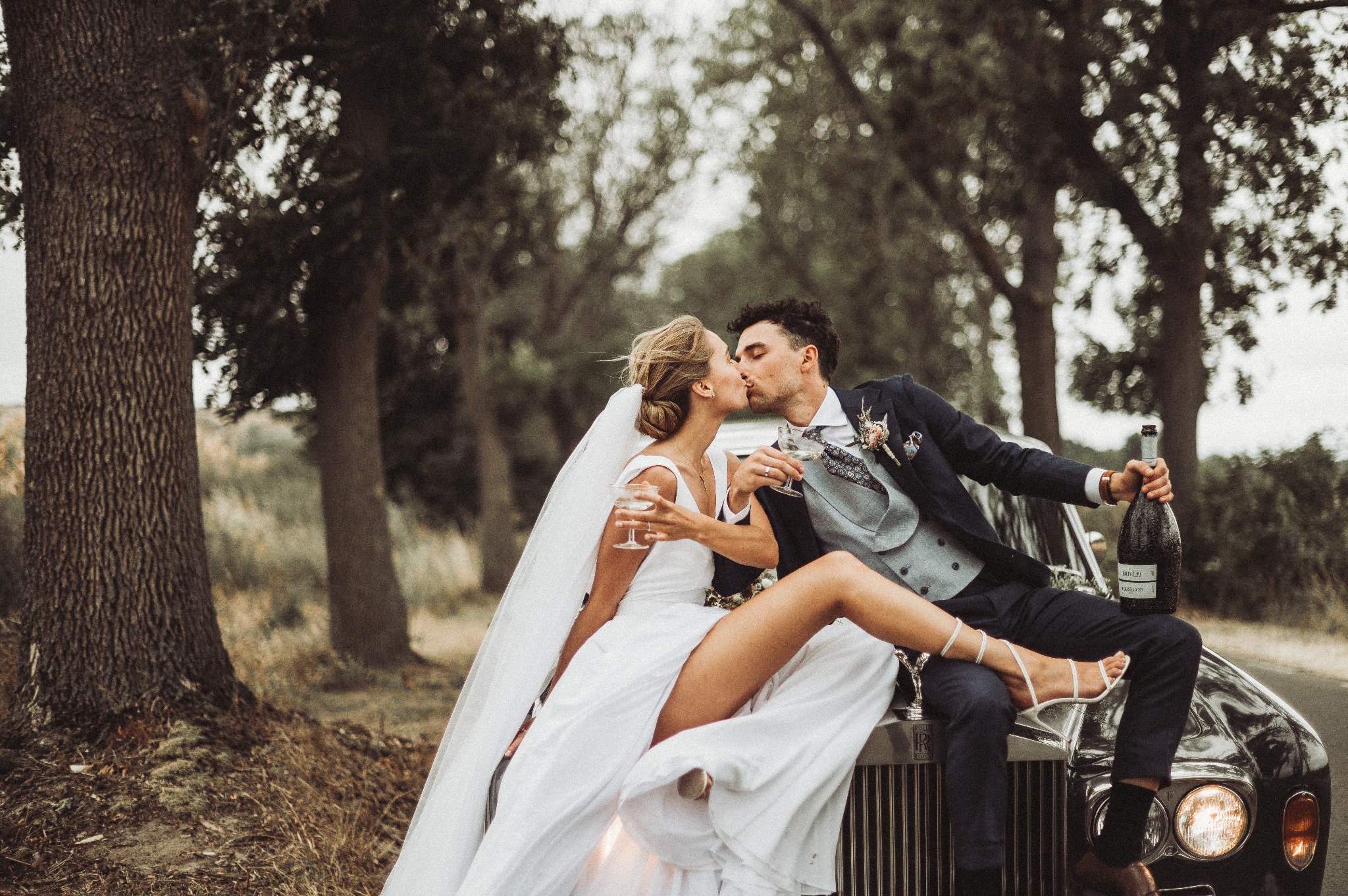 Para młoda w białej sukni i granatowym garniturze całuje się, siedząc na masce czarnego samochodu Rolls Royce, trzymając kieliszki z szampanem i butelkę w plenerze.