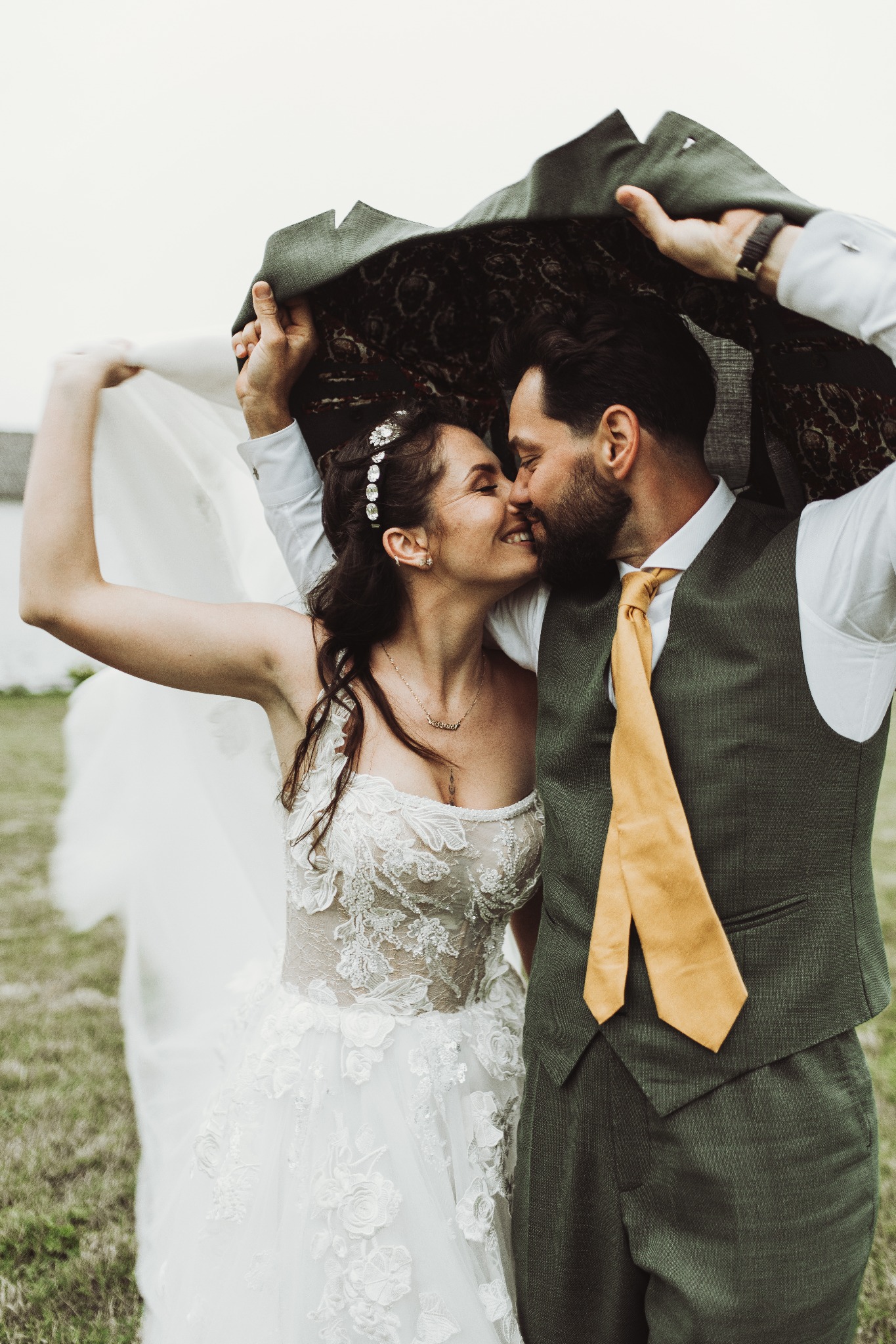 Pocałunek młodej pary pod marynarką w Baranowie: panna młoda w koronkowej sukni z tiulowym welonem, pan młody w zielonej kamizelce i żółtym krawacie.