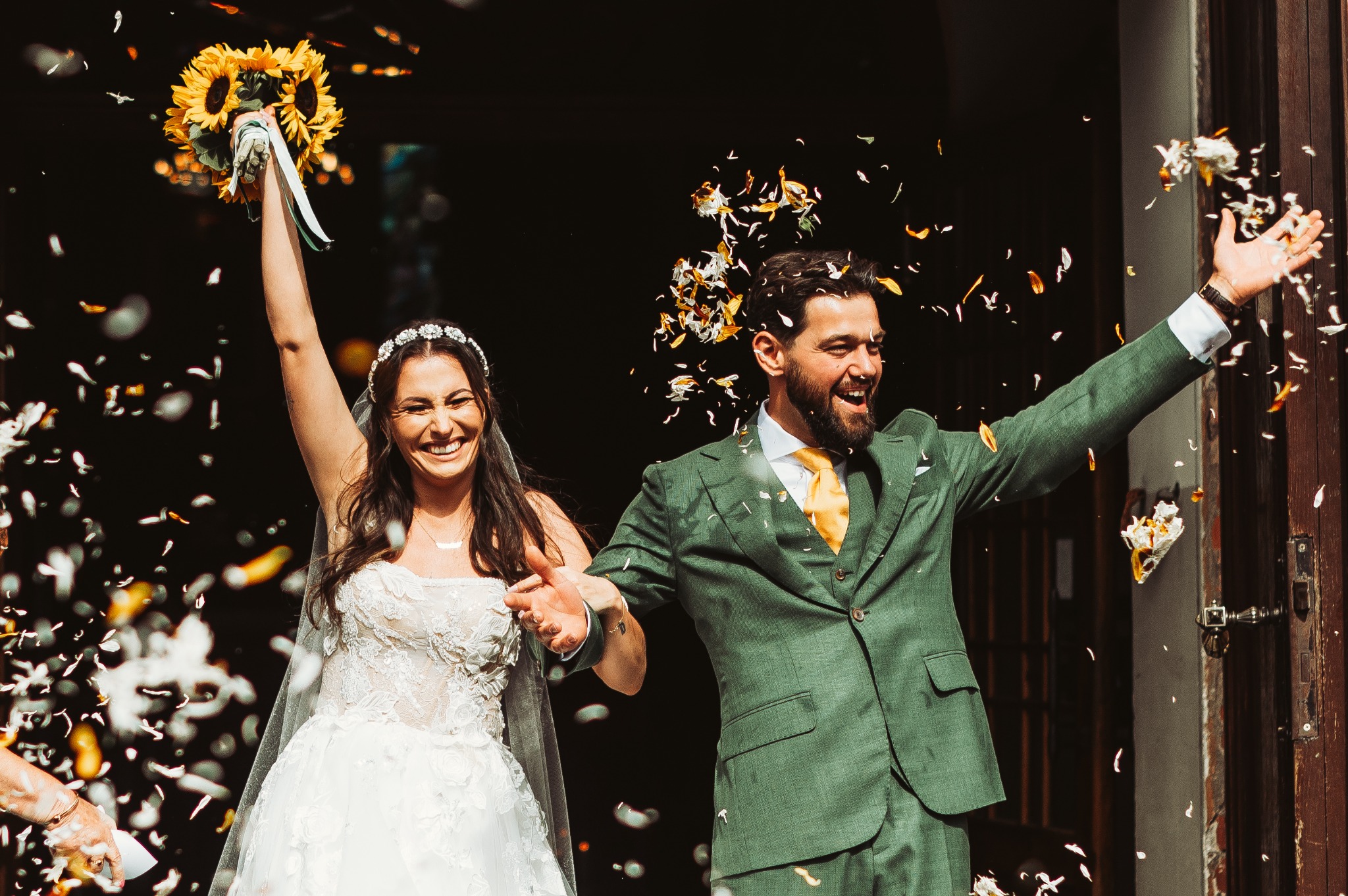 Uśmiechnięta para młoda wychodzi z kościoła obsypana konfetti, panna młoda z bukietem słoneczników w uniesionej ręce, pan młody w zielonym garniturze z żółtym krawatem.