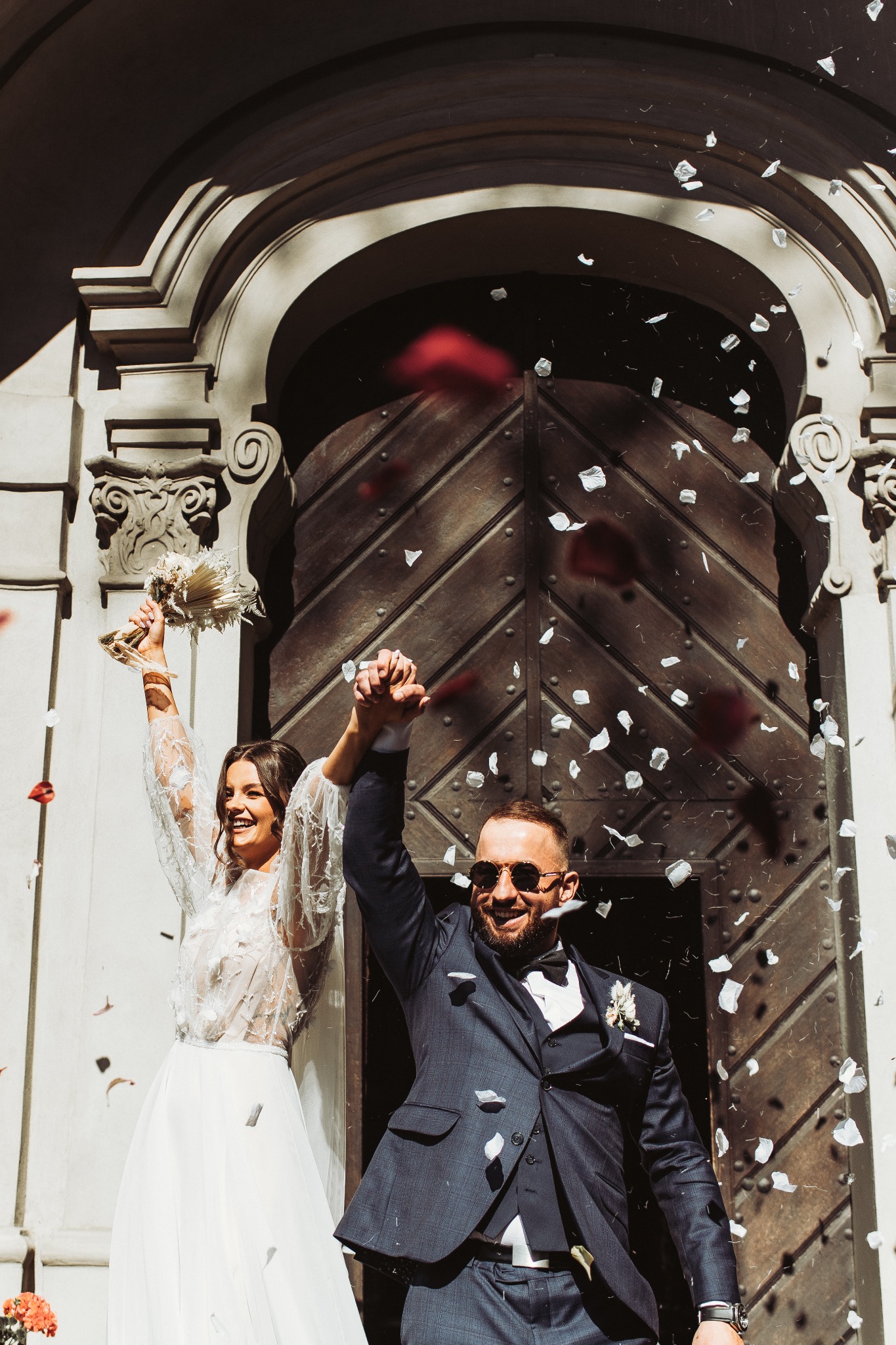 Uśmiechnięta para młoda wychodzi z kościoła, obsypywana płatkami kwiatów; panna młoda w koronkowej sukni, pan młody w granatowym garniturze i okularach przeciwsłonecznych.