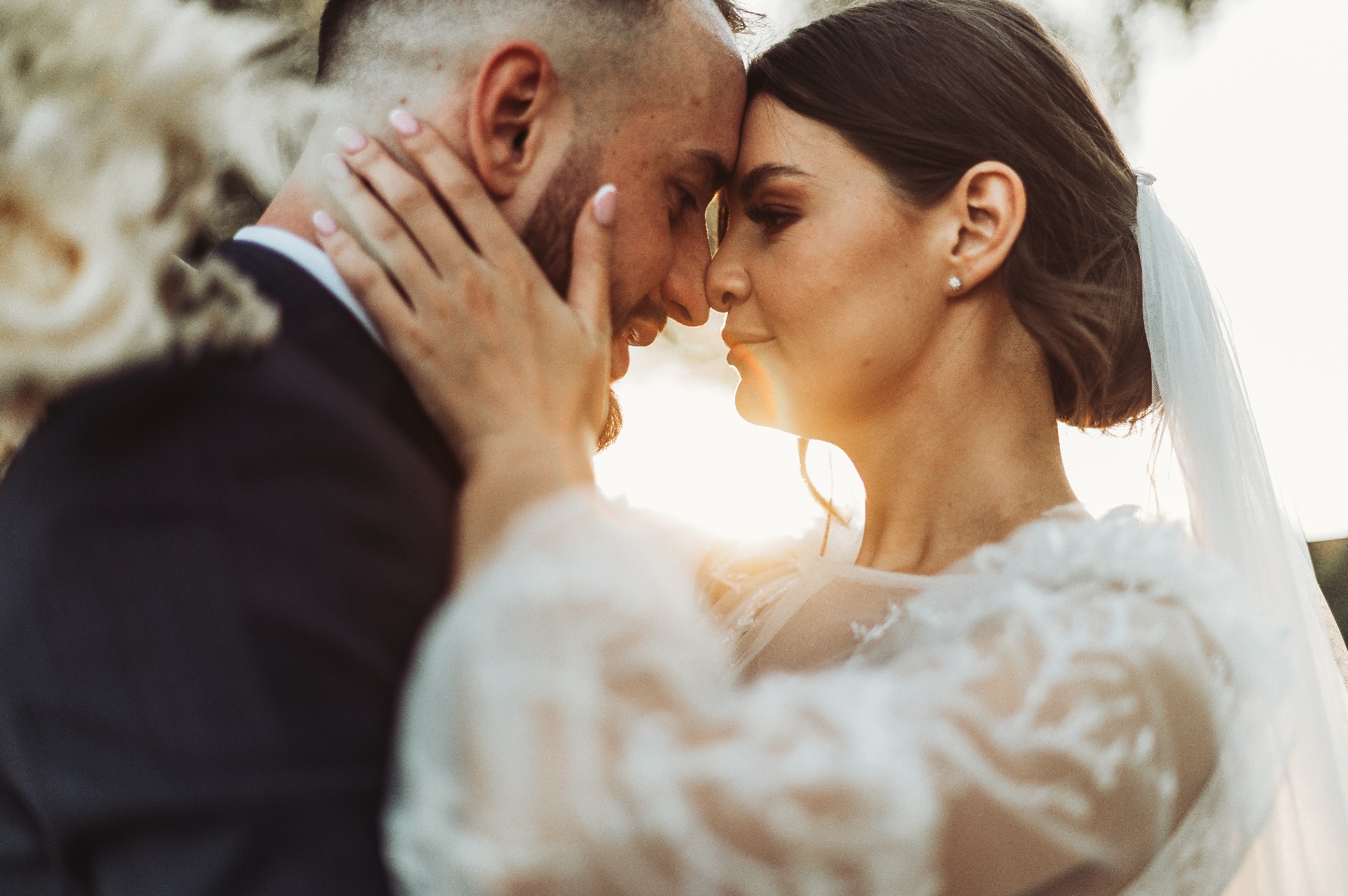 Para młoda, zbliżenie twarzy, delikatne światło słoneczne przebija przez welon, on w ciemnym garniturze, ona w koronkowej sukni, intymny moment.