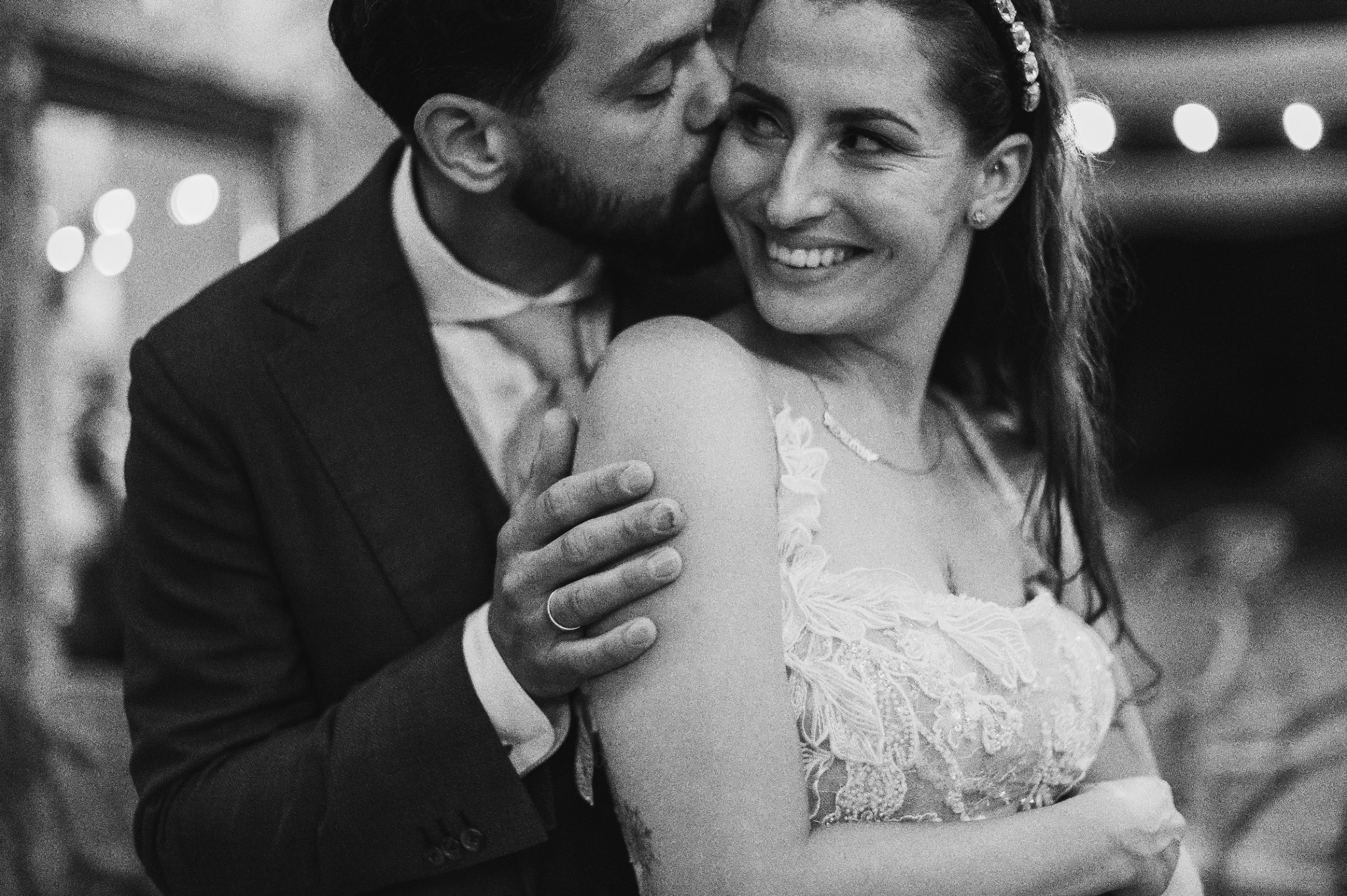 Czarno-białe zbliżenie na uśmiechniętą pannę młodą w koronkowej sukni, obejmowaną przez pana młodego w garniturze, który całuje ją w policzek; widoczne światła w tle.