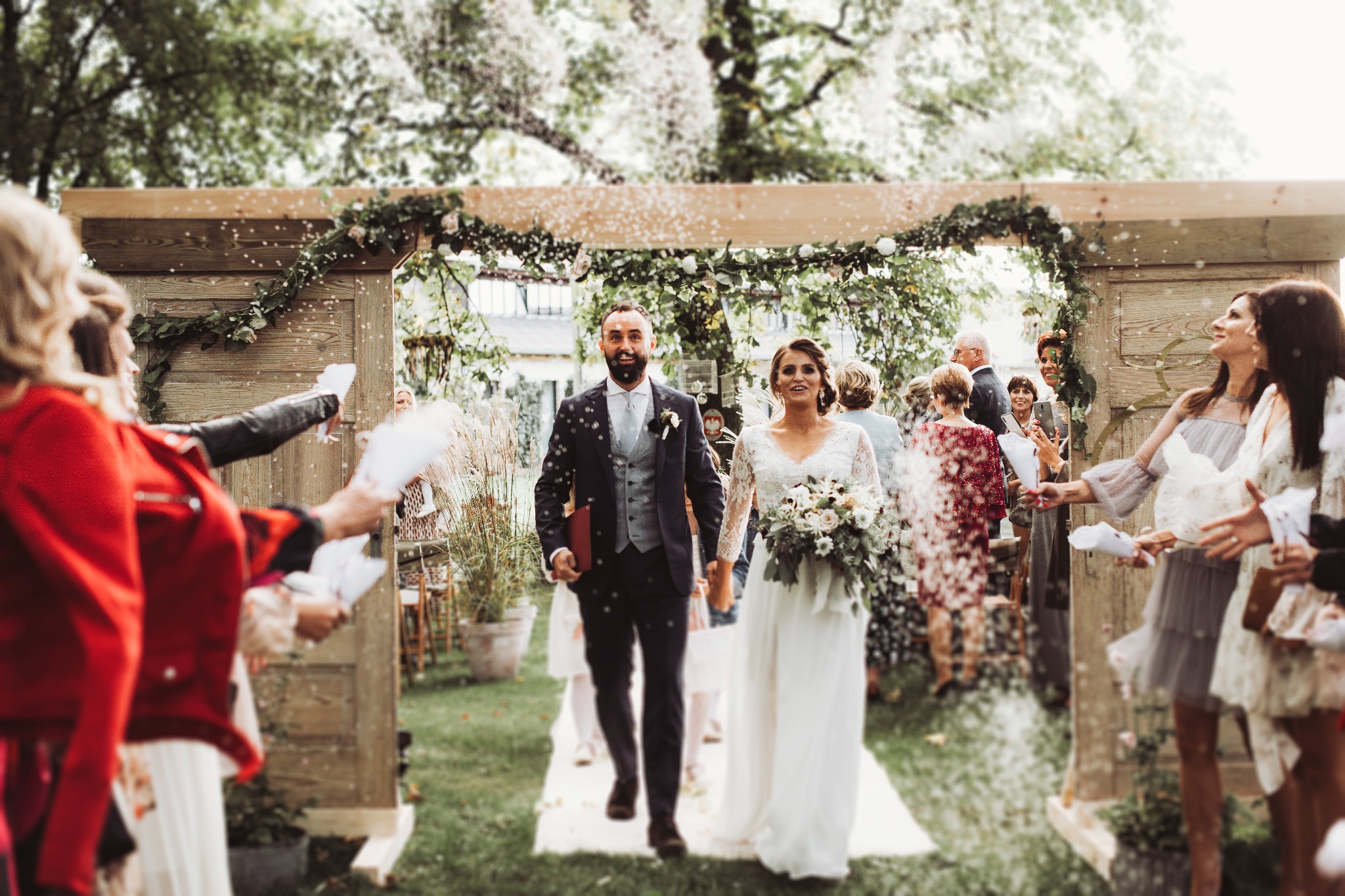 Para młoda, on w granatowym garniturze z szarą kamizelką, ona w białej sukni z koronkowymi rękawami, wychodzi spod drewnianego łuku udekorowanego zielenią i kwiatami, obsypywana konfetti przez gości.