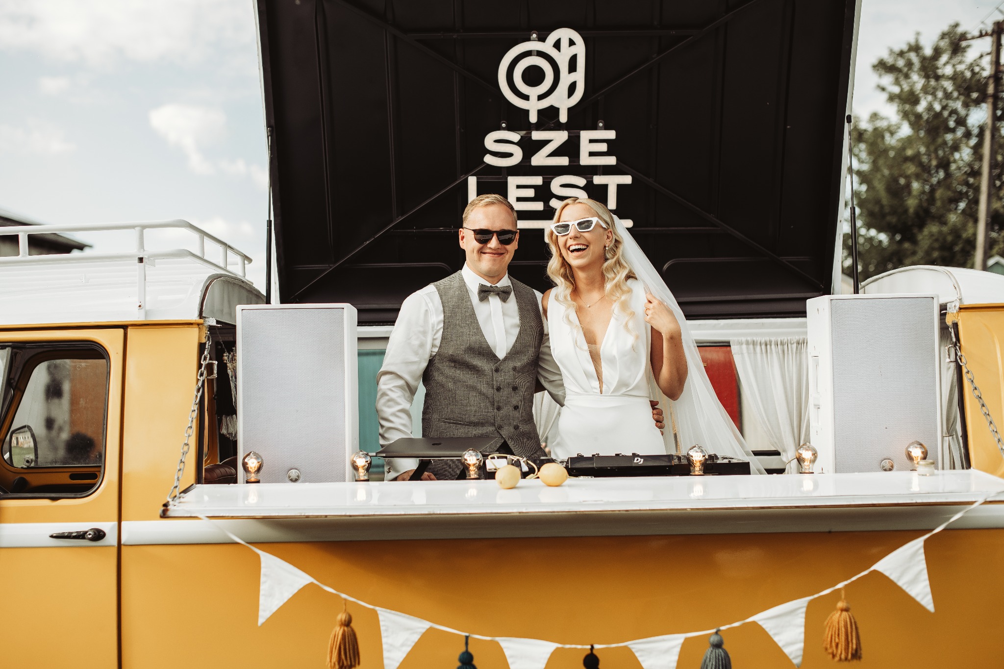 Uśmiechnięta para młoda w białych okularach pozuje za konsoletą DJ-ską umieszczoną w przerobionym żółtym vanie z napisem 'SZE LEST' w tle.