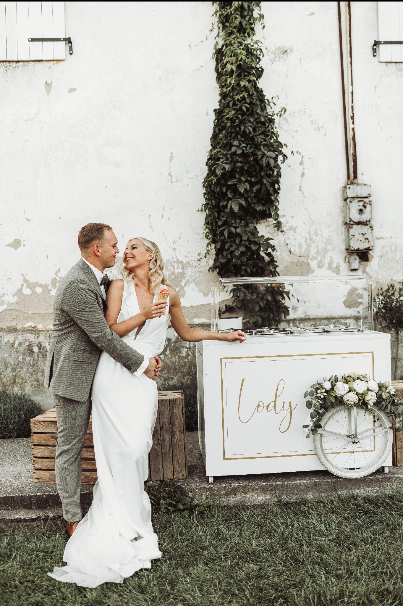 Para młoda w objęciach przed wózkiem z lodami z napisem 'Lody' i dekoracją kwiatową na tle białej ściany porośniętej bluszczem.