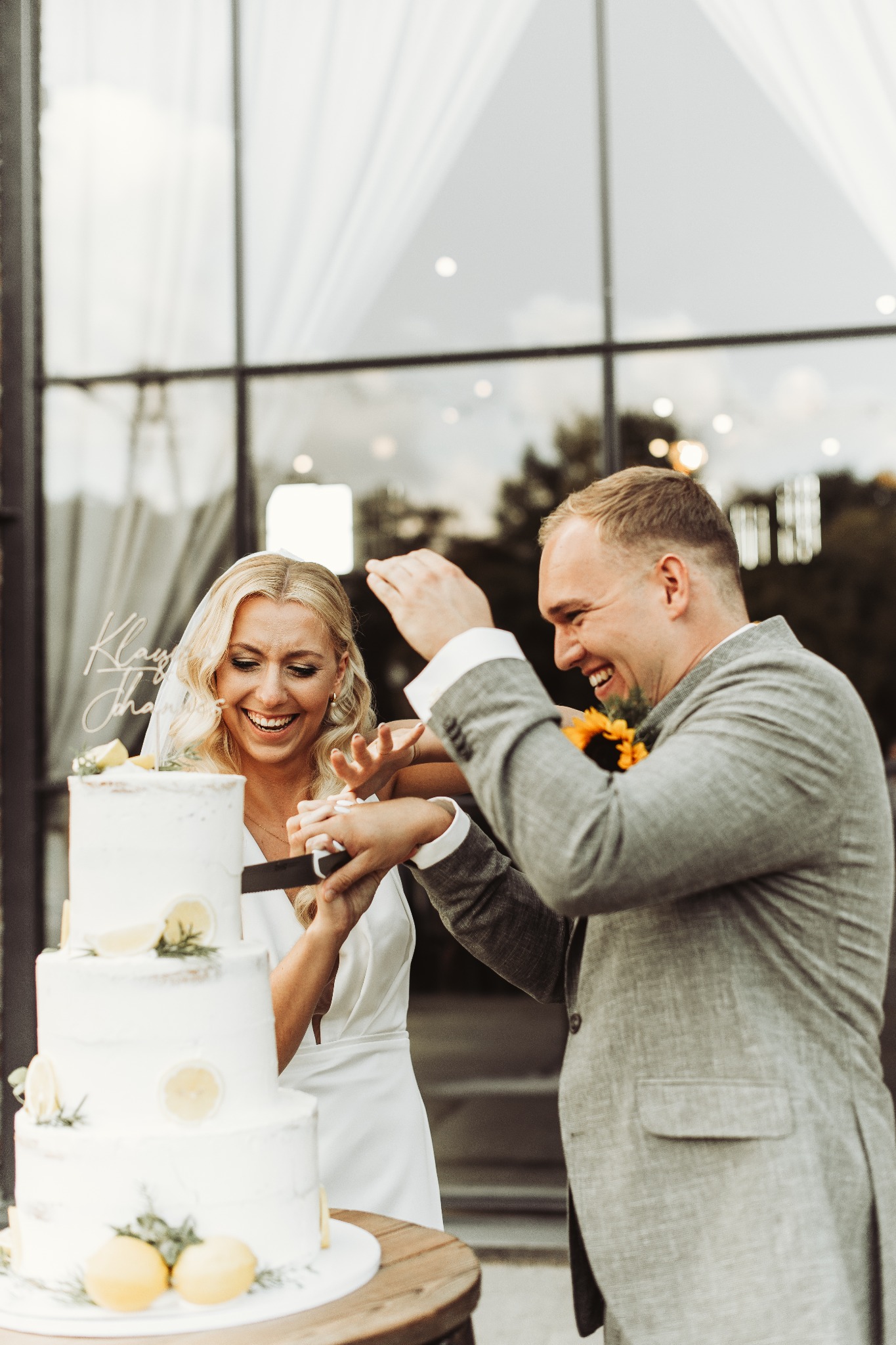 Uśmiechnięta para młoda kroi tort weselny udekorowany cytrynami i napisem z imionami, na tle przeszklonej ściany.