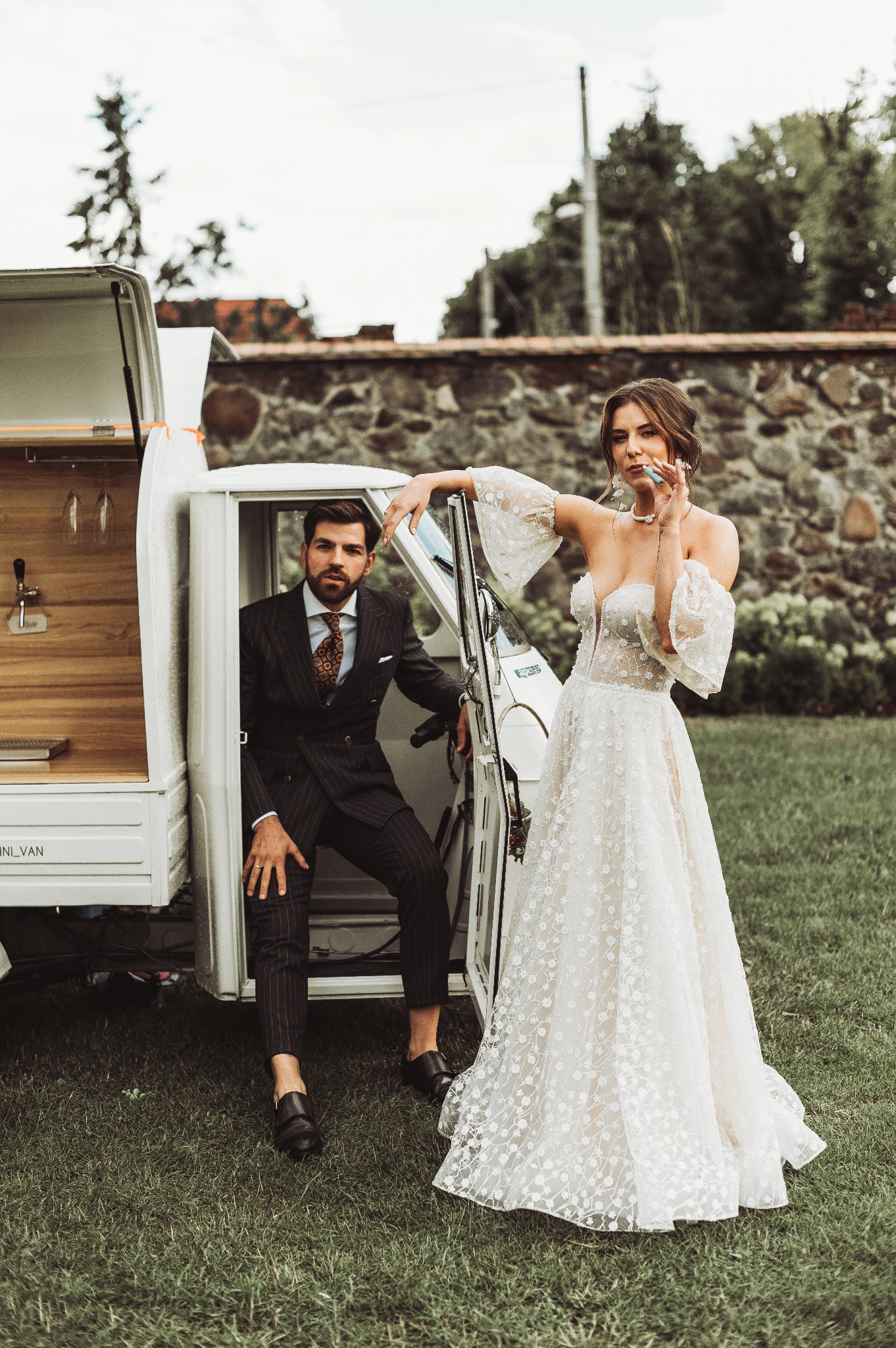 Elegancka para młoda pozuje przy przerobionym samochodzie dostawczym z barem, on w garniturze w prążki, ona w koronkowej sukni ślubnej z bufiastymi rękawami, na tle kamiennego muru i zielonego...