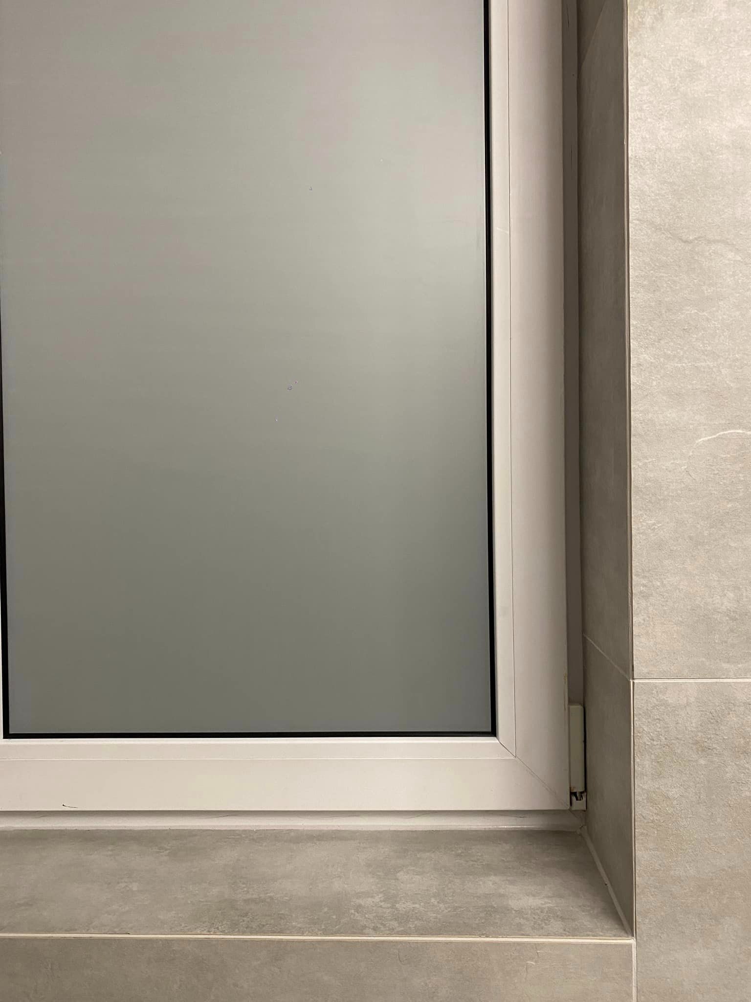 Zamknięte białe okno z matową szybą w łazience wyłożonej szarymi płytkami.