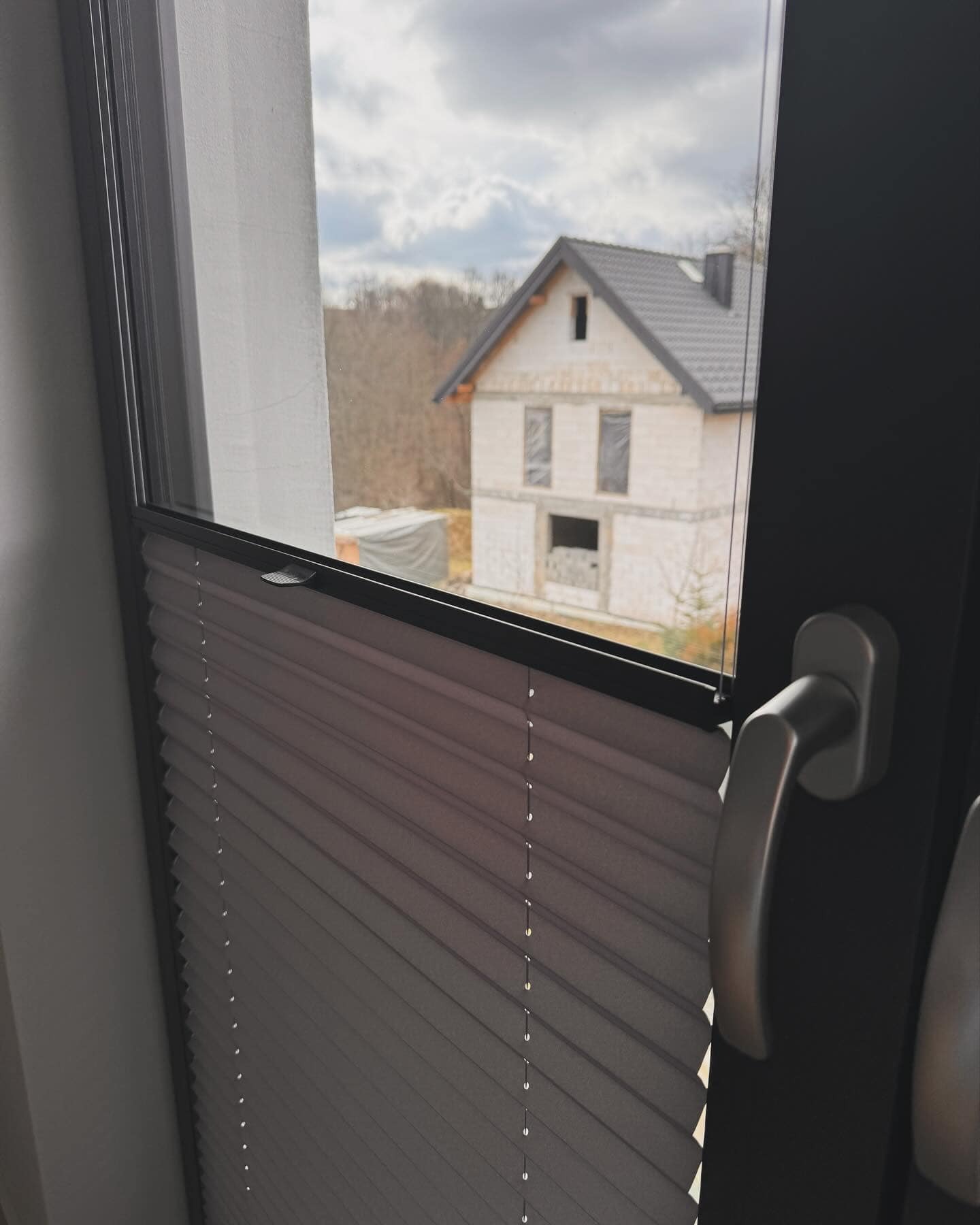 Szare plisowane osłony okienne w oknie z czarną ramą, widok na budynek w stanie surowym z zewnątrz.