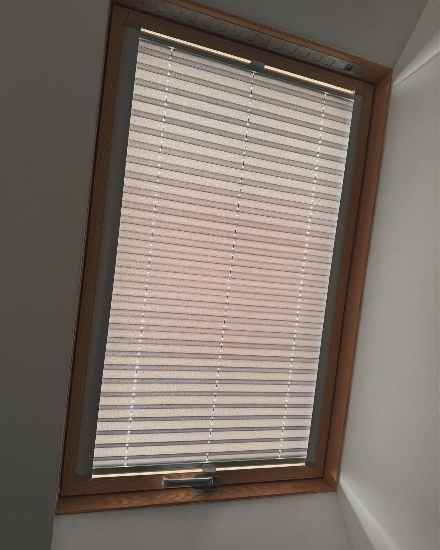 Beżowe plisowane rolety w oknie dachowym z drewnianą ramą, widoczny mechanizm sterujący.