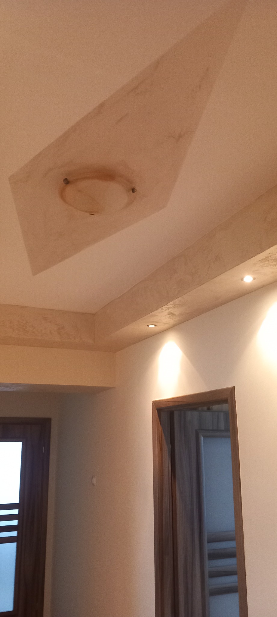 Wykończony sufit z dekoracyjną gładzią w odcieniach beżu, oświetlenie punktowe i lampa sufitowa, widok na fragment ściany z drzwiami.