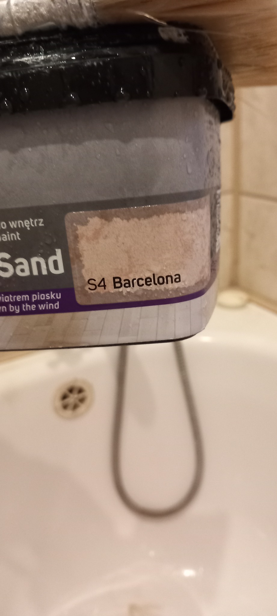 Puszka farby do wnętrz w odcieniu piaskowym S4 Barcelona, z widocznymi kroplami wody na wieczku, umieszczona nad białą umywalką.