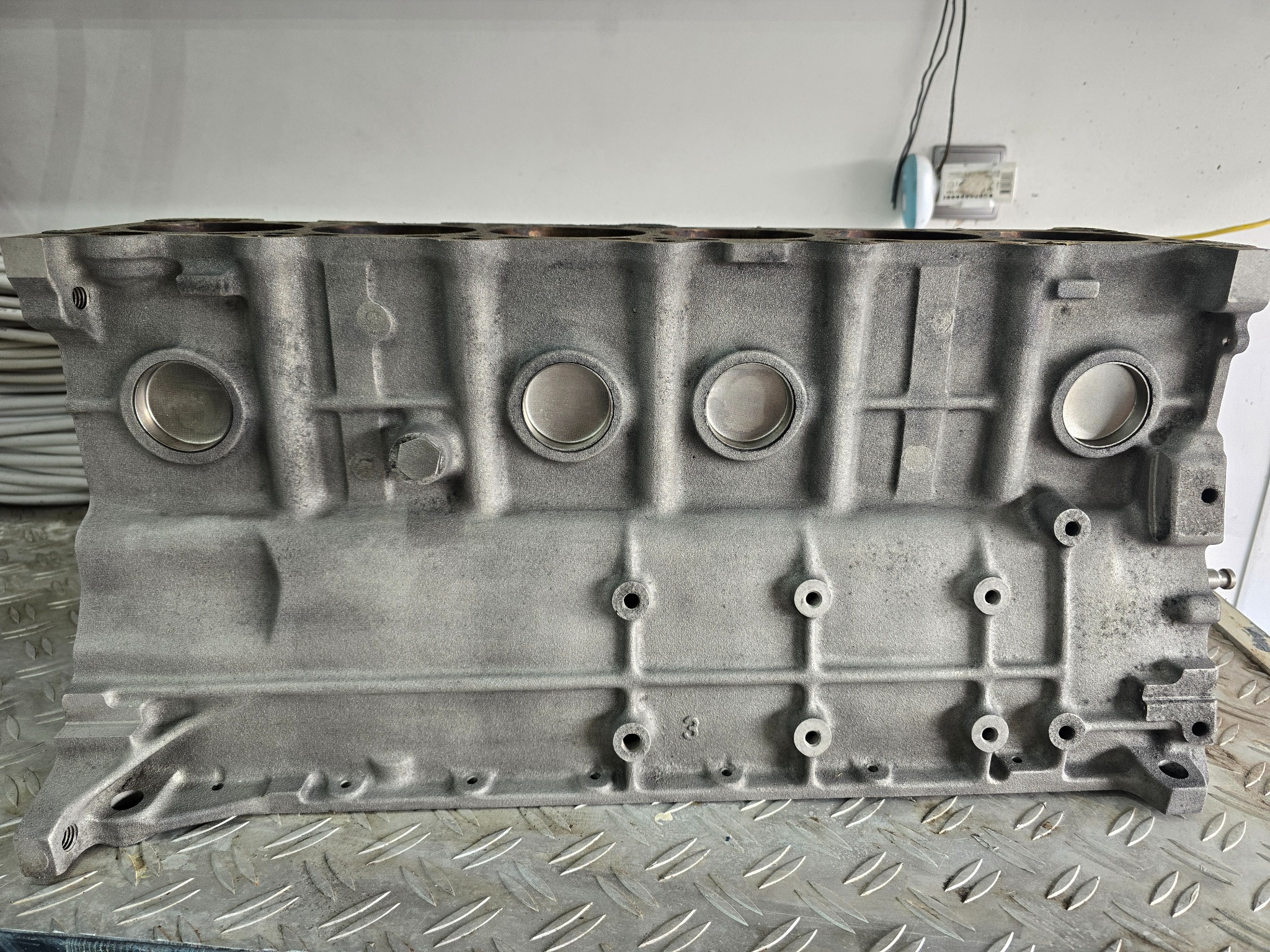 Szlifowany blok silnika samochodowego z widocznymi otworami na cylindry i śruby, umieszczony na metalowej powierzchni o wzorze rombów.