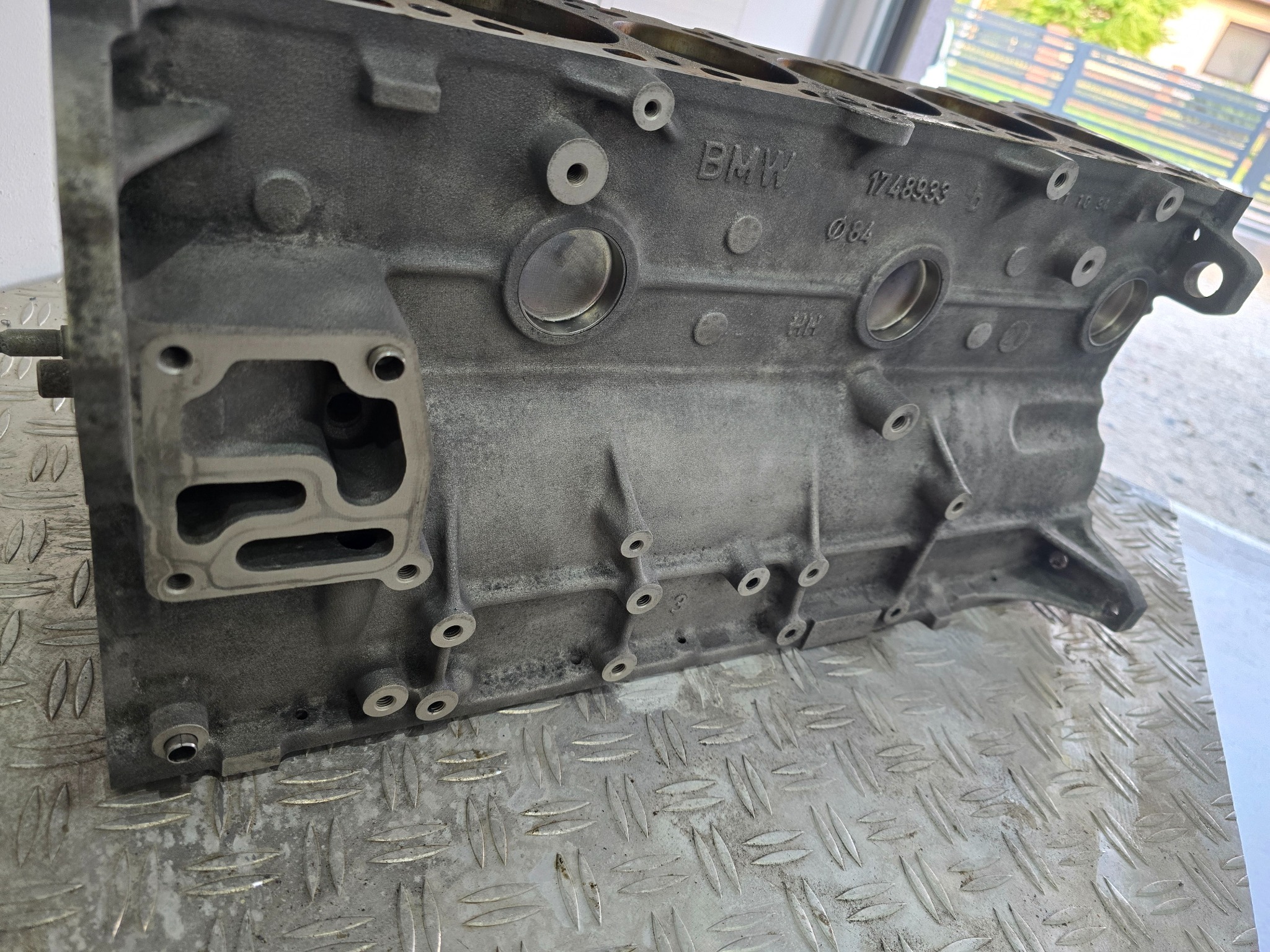 Blok silnika BMW na metalowej powierzchni o strukturze rombów, widoczne otwory i oznaczenia fabryczne.