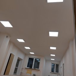 WILPRO Adam Wilk - Wnętrze w trakcie remontu z rzędem kwadratowych paneli LED w suficie, widoczne worki z zaprawą i narzędzia malarskie przy ścianach.