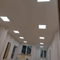 Wnętrze w trakcie remontu z rzędem kwadratowych paneli LED w suficie, widoczne worki z zaprawą i narzędzia malarskie przy ścianach.
