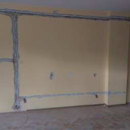 WILPRO Adam Wilk - Wnętrze w trakcie remontu z widocznymi przewodami elektrycznymi w białych rurkach ułożonymi wzdłuż ścian, częściowo zamaskowanymi gipsem, z widocznymi puszkami instalacyjnymi.