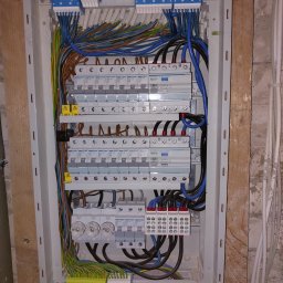 WILPRO Adam Wilk - Otwarta skrzynka elektryczna z zainstalowanymi bezpiecznikami, przewodami o różnych kolorach i złączkami, zamontowana na ścianie z surowego drewna i betonu.