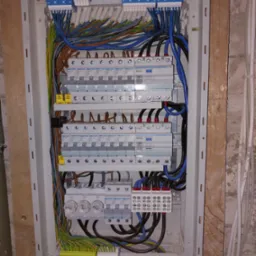 Otwarta skrzynka elektryczna z zainstalowanymi bezpiecznikami, przewodami o różnych kolorach i złączkami, zamontowana na ścianie z surowego drewna i betonu.