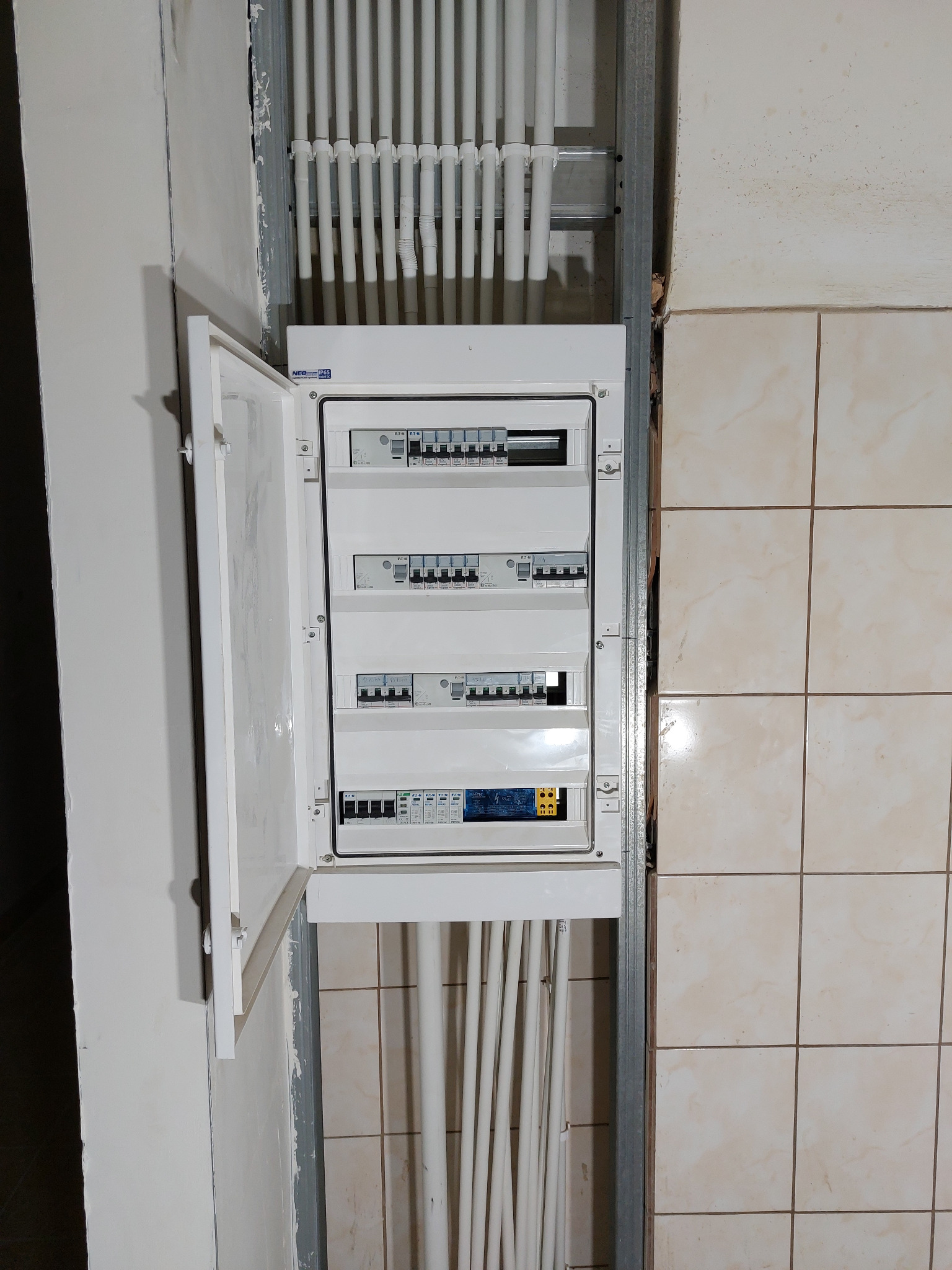 Otwarta skrzynka bezpiecznikowa z widocznymi bezpiecznikami i przewodami w ścianie z białymi rurkami instalacyjnymi.