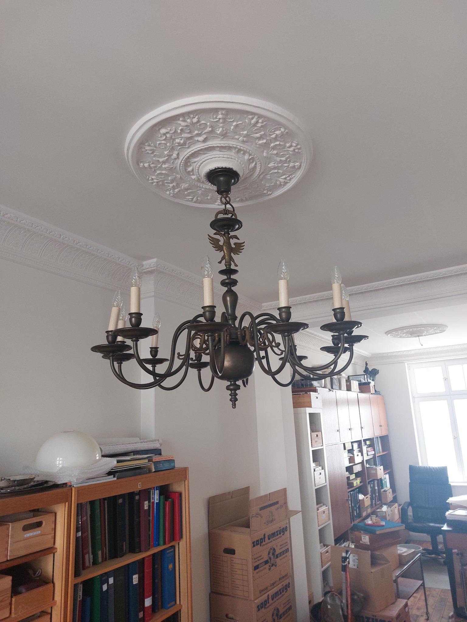 Antyczny, wieloramienny żyrandol z brązu z ozdobnymi świecznikami i figurką orła, zawieszony na suficie z dekoracyjną rozetą.