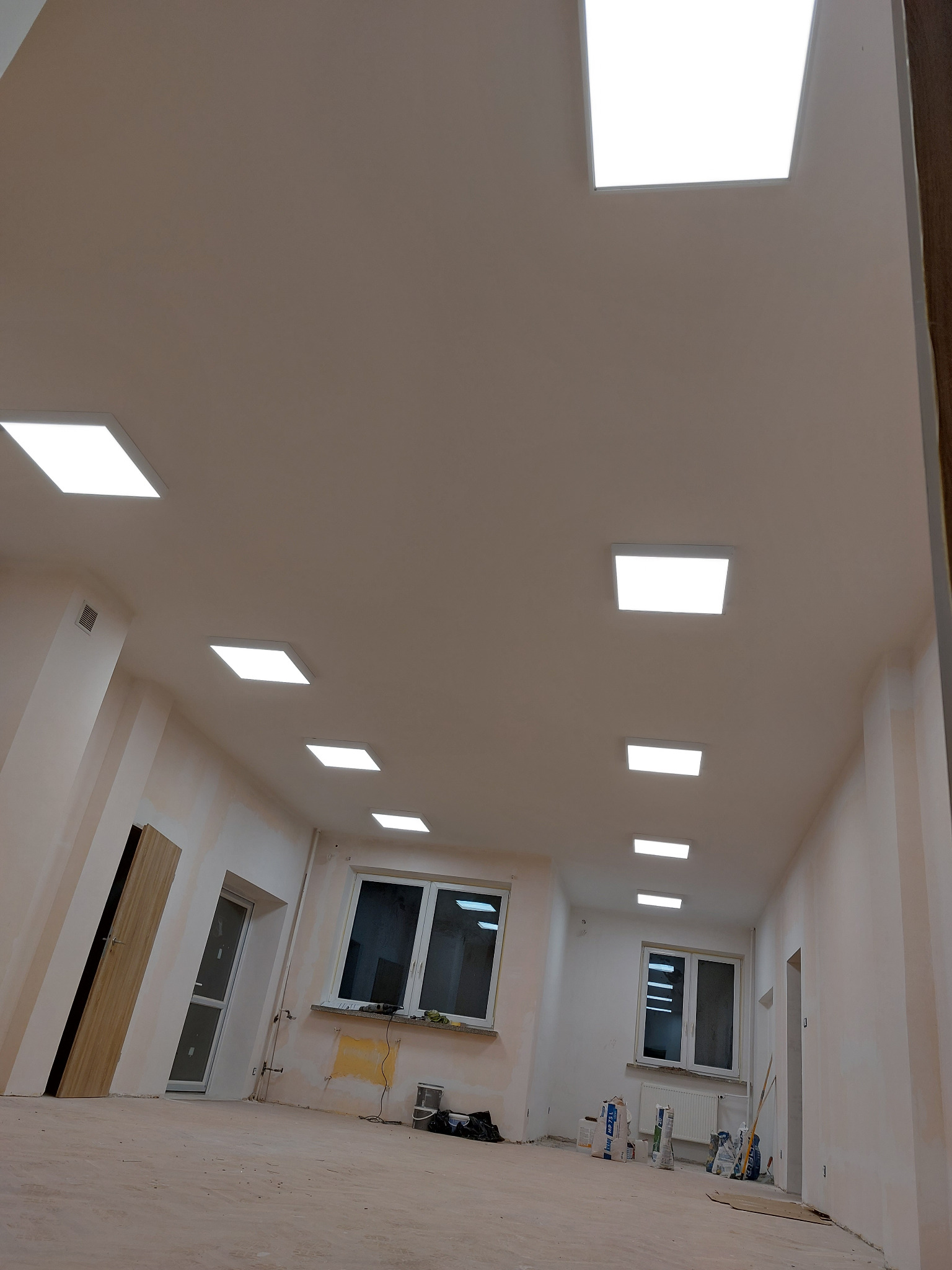 Wnętrze w trakcie remontu z rzędem kwadratowych paneli LED w suficie, widoczne worki z zaprawą i narzędzia malarskie przy ścianach.