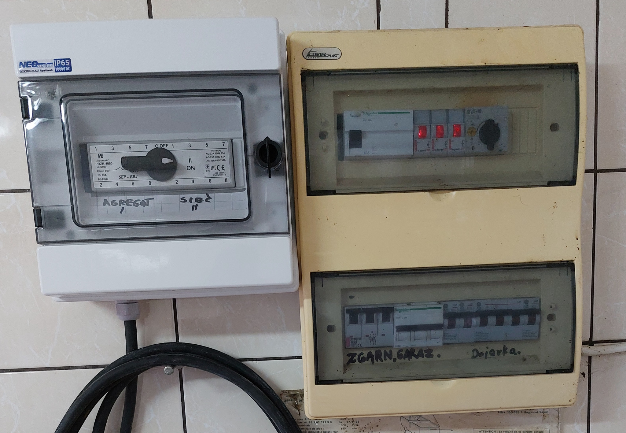 Dwa stare liczniki elektryczne obok siebie, jeden nowszy biały z przełącznikiem agregat/sieć, drugi starszy żółty z napisem 'ZGarn, Garaz' i 'Dojarka' na tle kafelków.