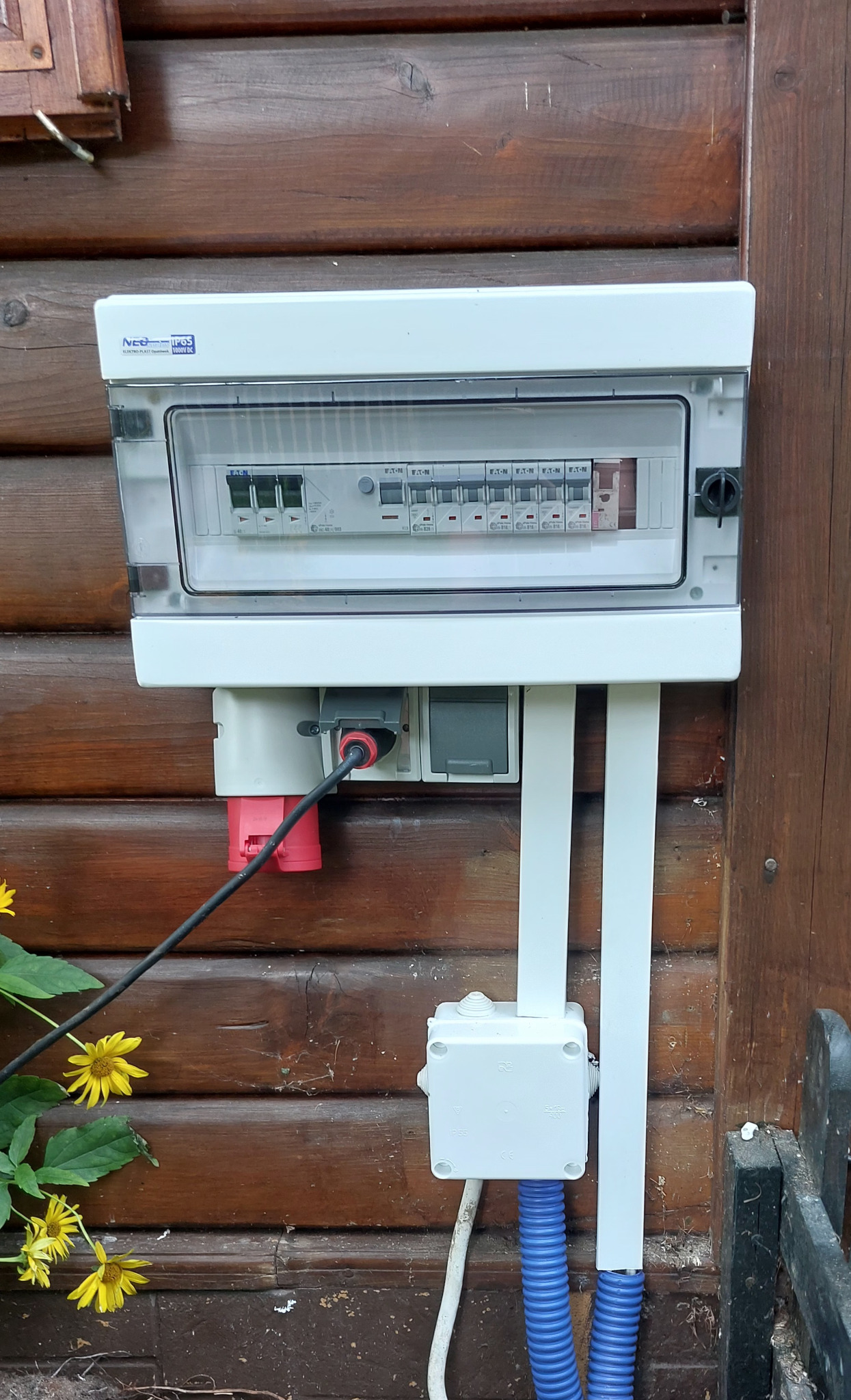 Zewnętrzna skrzynka elektryczna z zabezpieczeniami i gniazdami na drewnianej elewacji, widoczne kable w białych i niebieskich peszlach, obok kwitnące żółte kwiaty.